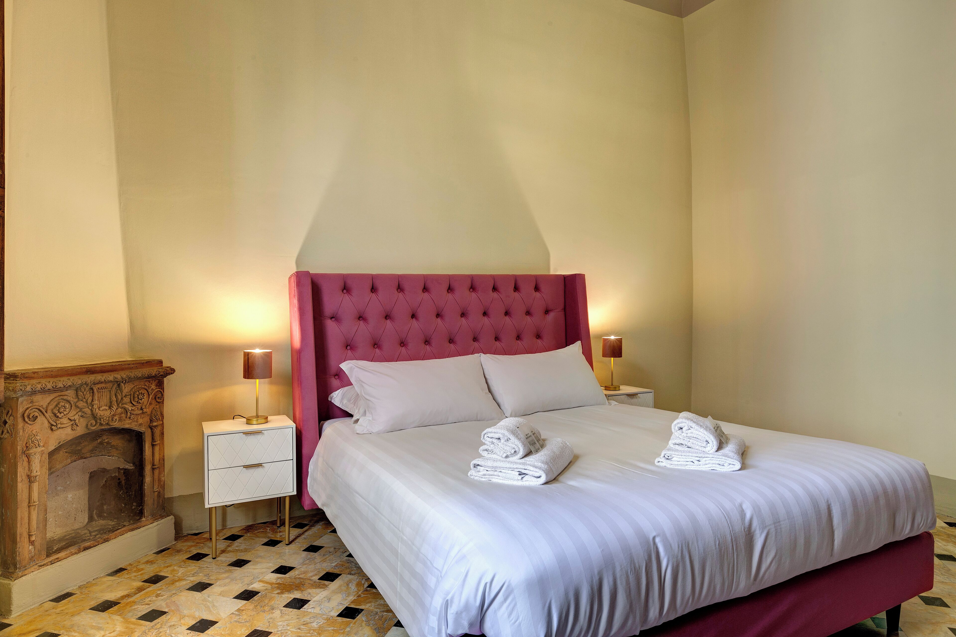 Un lit bien fait, avec du linge de lit blanc et deux serviettes pliées, une tête de lit rembourrée de couleur rouge, une petite table de chevet avec une lampe et une cheminée avec un sol carrelé.