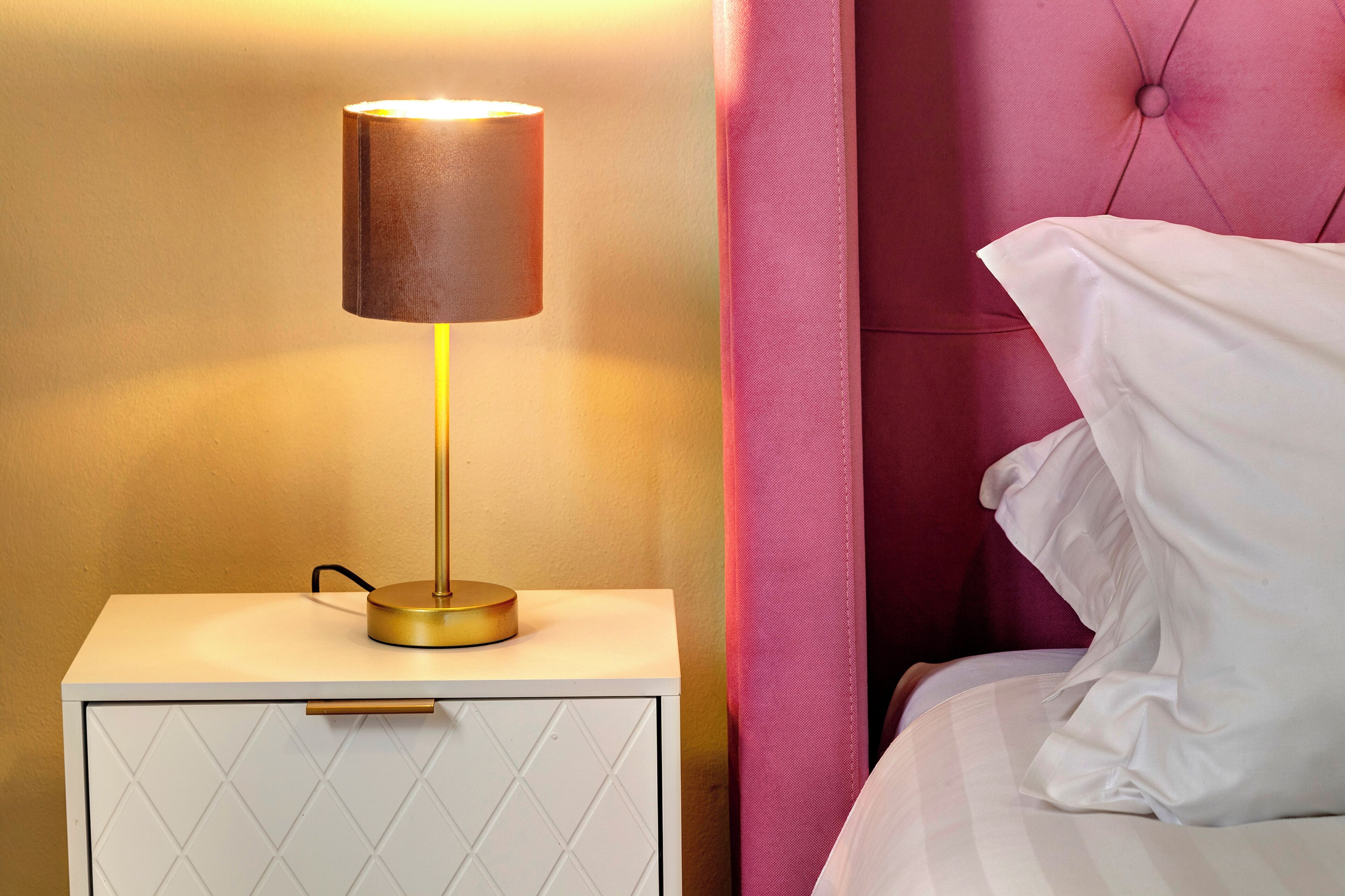 Une table de chevet avec une lampe, un lit avec une tête de lit rose et un oreiller blanc.