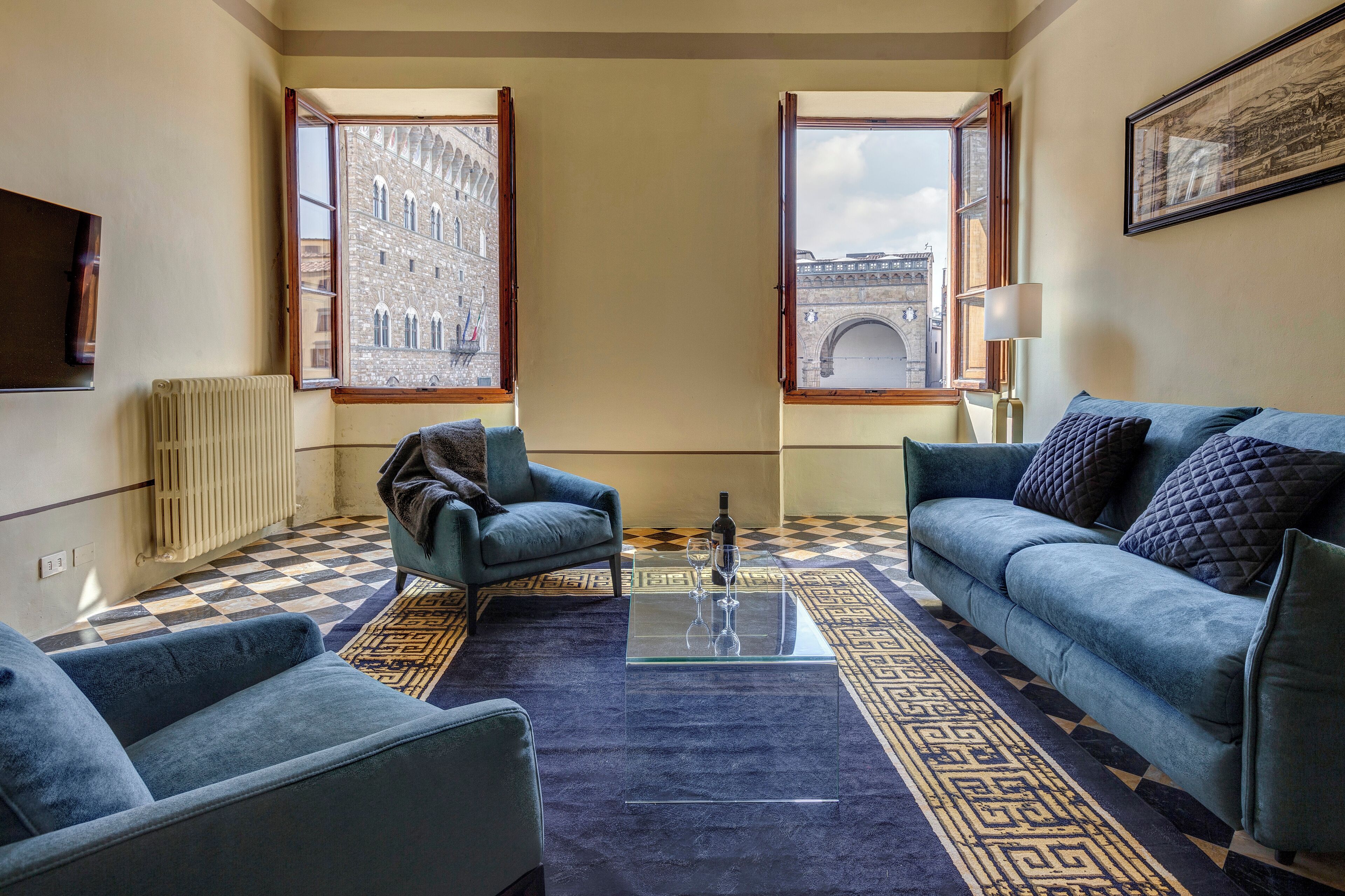 Ein Wohnzimmer mit Schachbrettmuster-Boden, einem Sofa, einem Sessel, einem Glastisch und zwei Fenstern mit Blick auf ein Gebäude und einen Bogen.