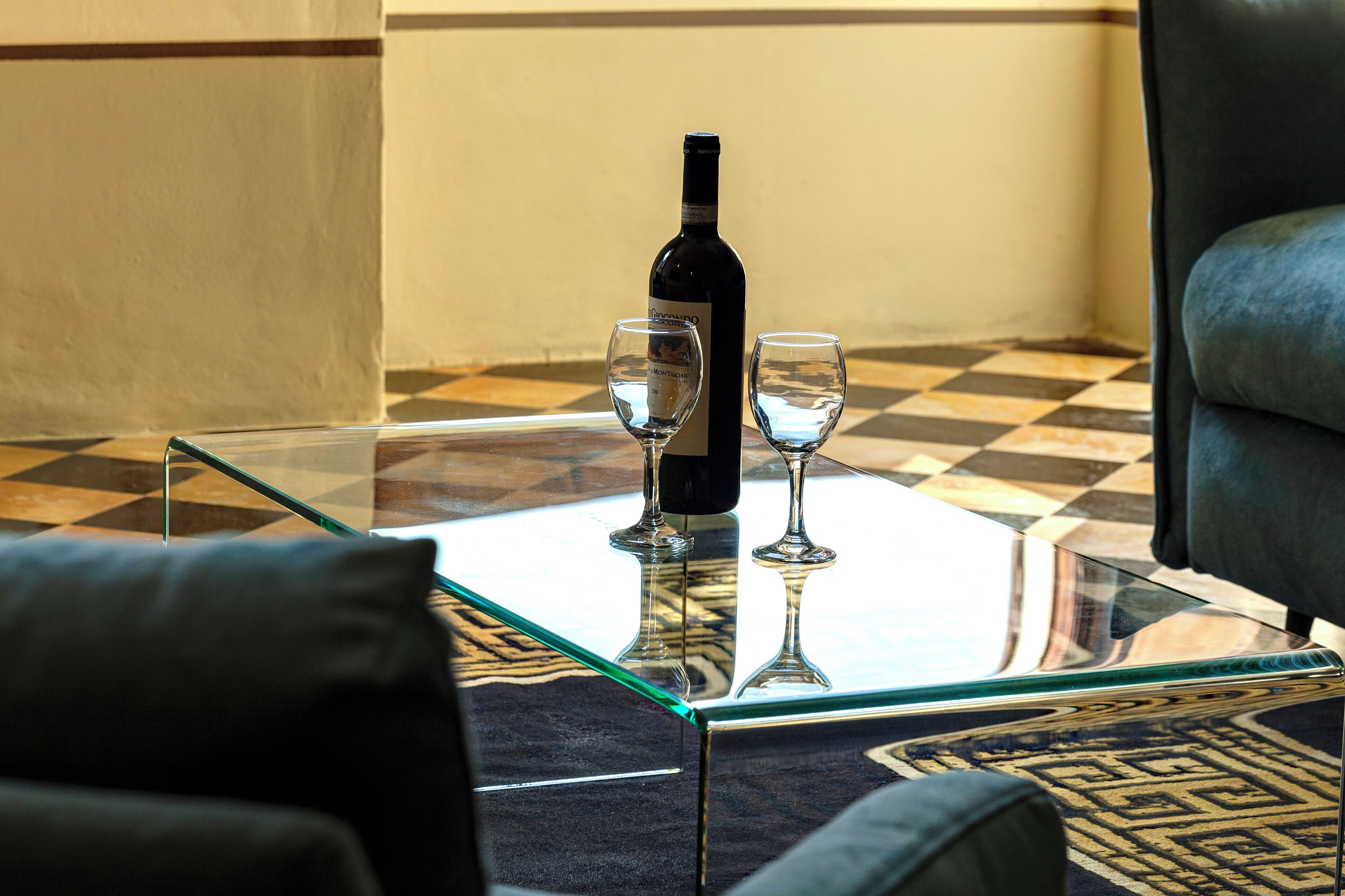 Une table en verre sur laquelle sont posés une bouteille de vin et deux verres, le tout entouré d’un sol à carreaux et d’un tapis à motifs.
