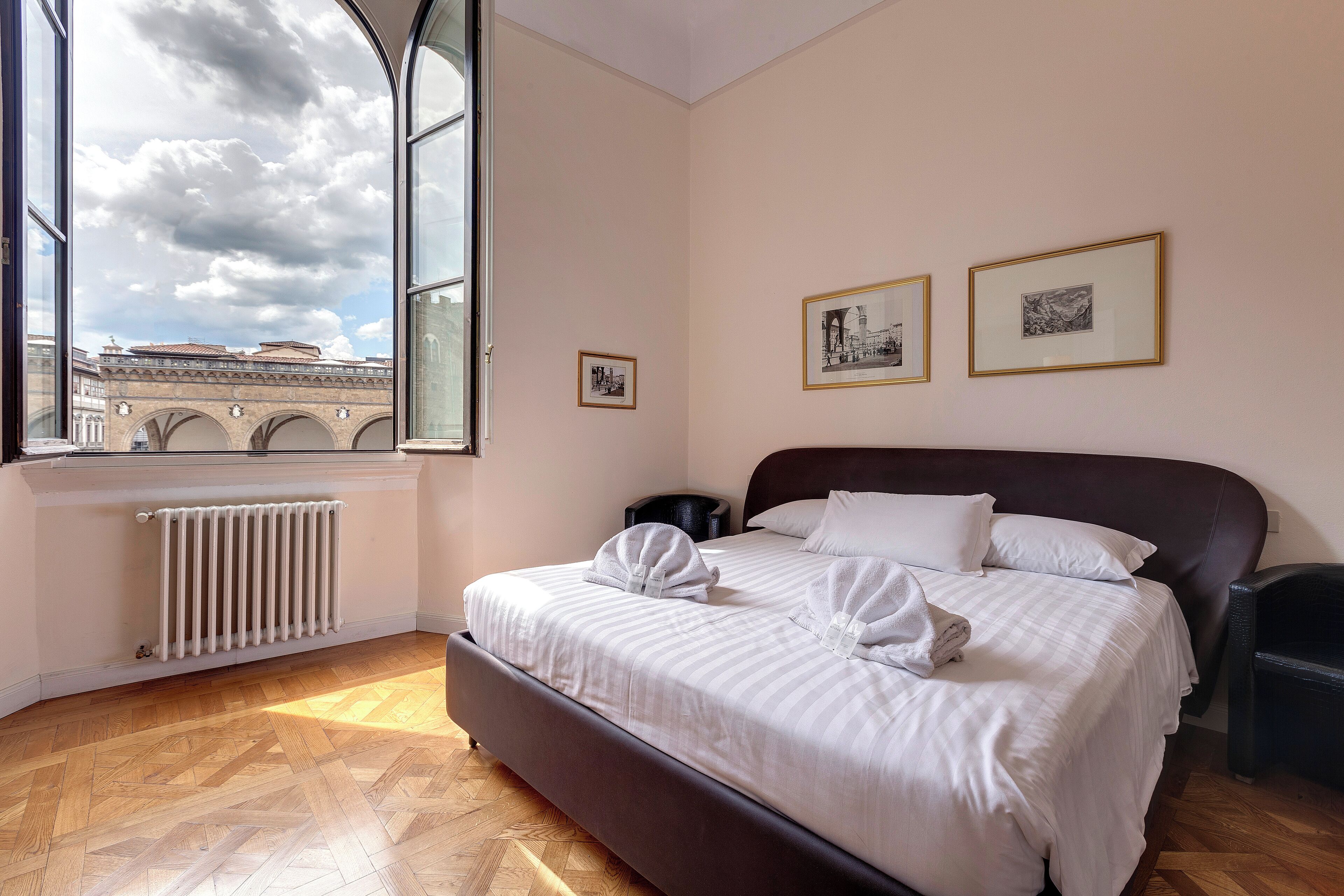 Ein Schlafzimmer mit großem Fenster, Heizkörper, einem Bett mit weißer Bettwäsche und zwei gerahmten Bildern an der Wand.