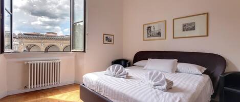 Ein Schlafzimmer mit großem Fenster, Heizkörper, einem Bett mit weißer Bettwäsche und zwei gerahmten Bildern an der Wand.