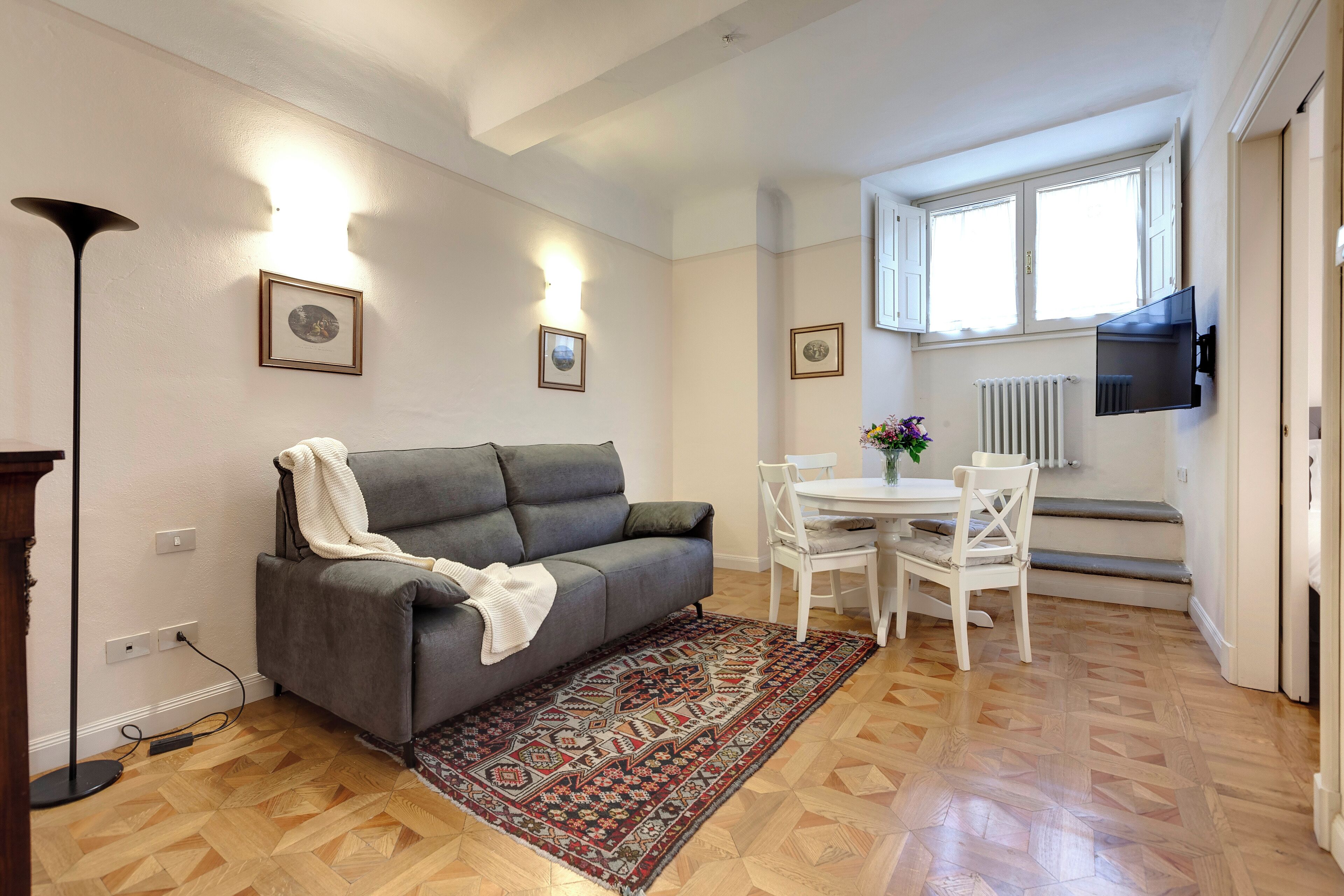 A living room with a grey sofa, a patterned rug, a white dining table with chairs, and a TV mounted on the wall.