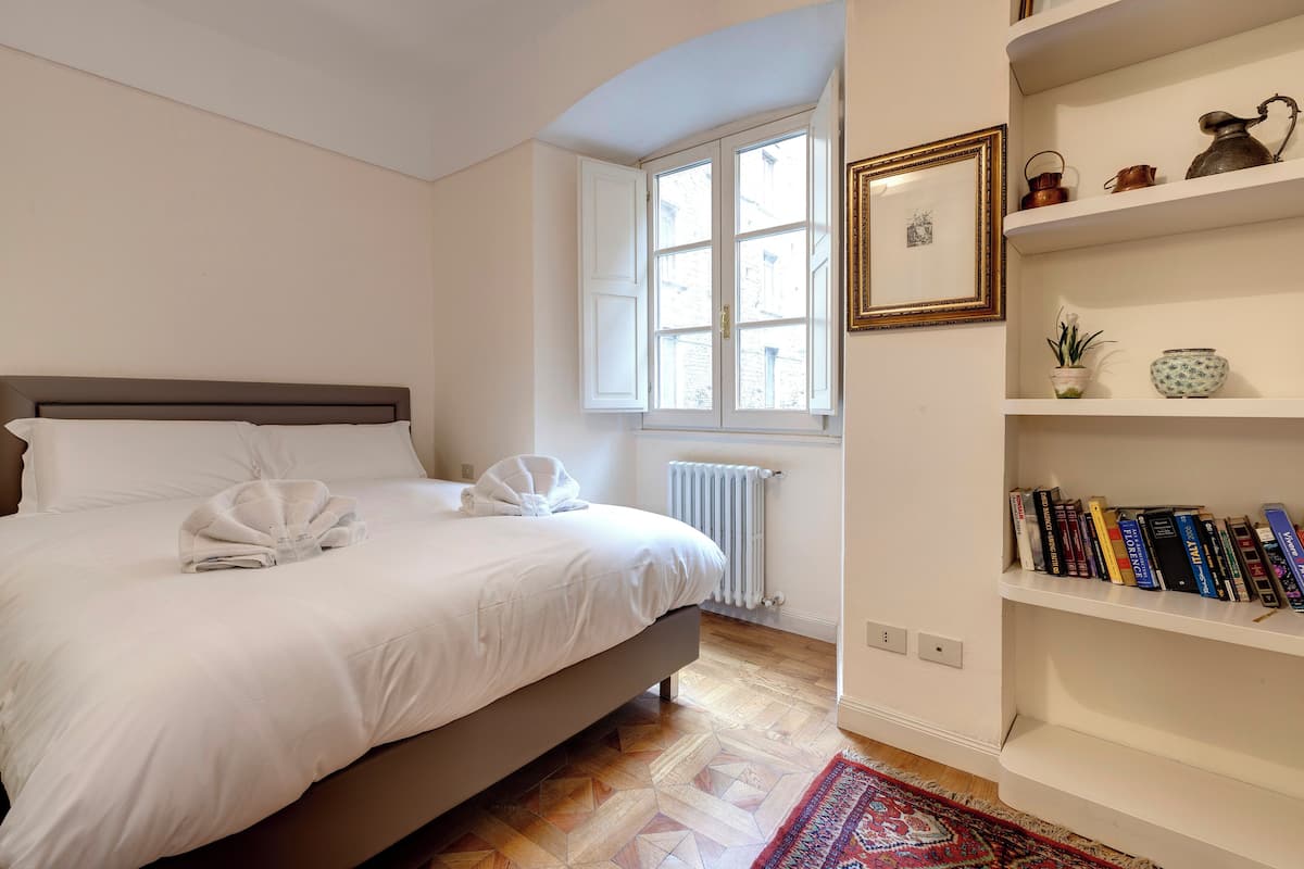 A bedroom with a bed, a radiator, a window, and a bookshelf.