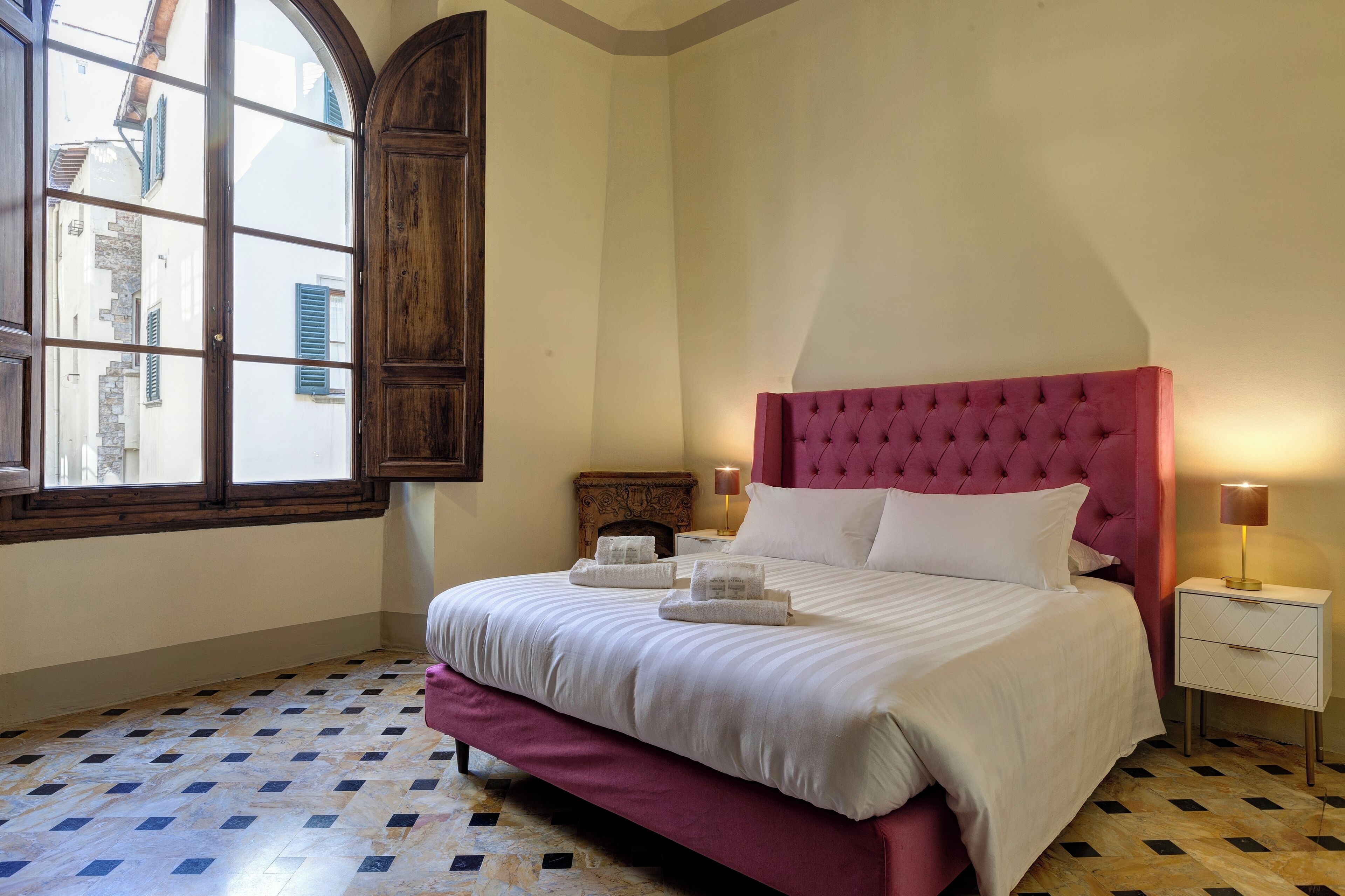 A bedroom with a large bed, a wooden wardrobe, a nightstand with a lamp, and a window with shutters.