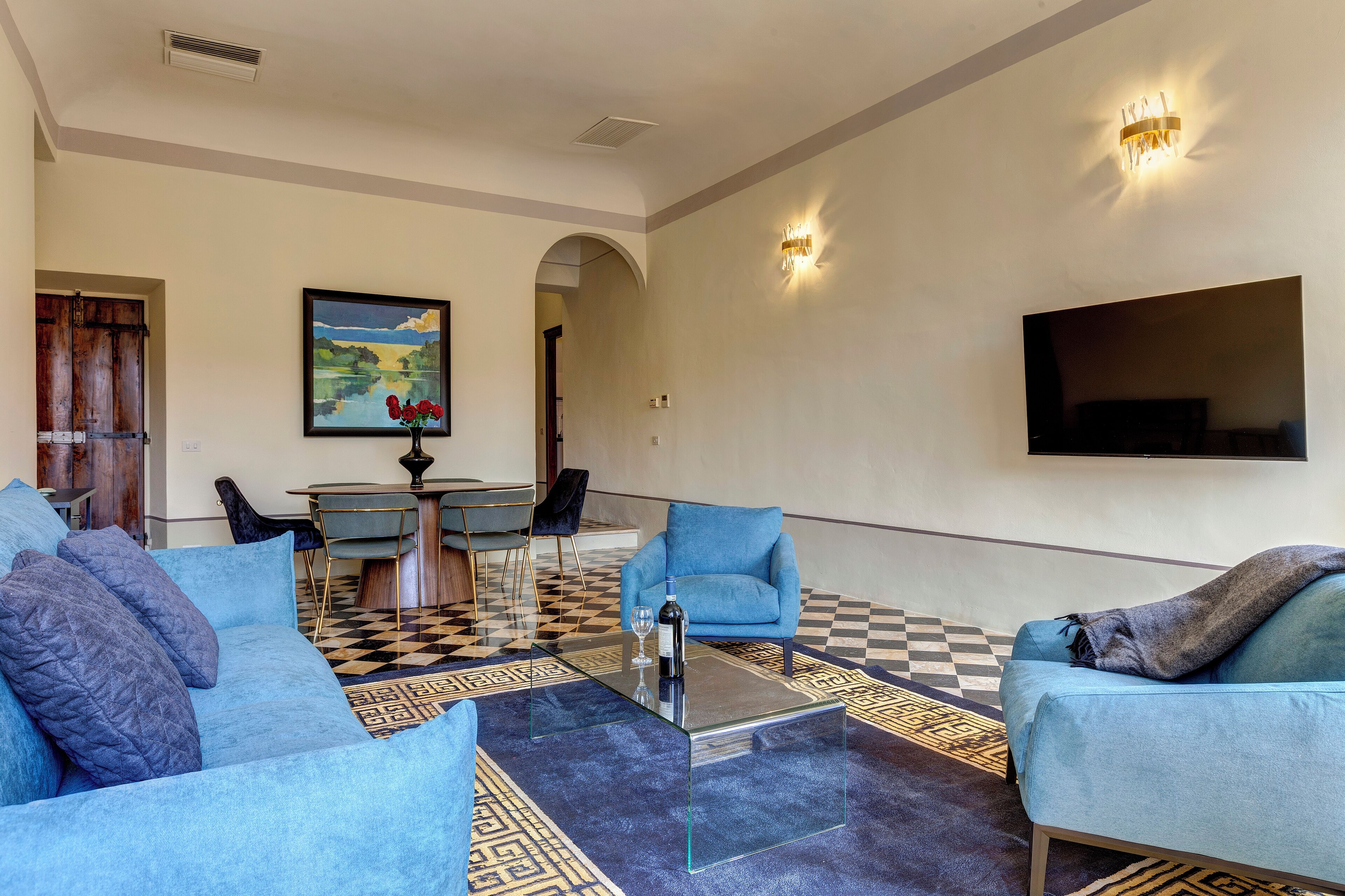 Een moderne woonkamer met een zwart-wit geruit vloerkleed, een glazen salontafel, blauwe banken en een schilderij aan de muur.
