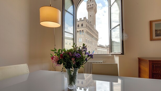 Una habitación con una mesa de comedor, un jarrón de flores, una lámpara y vista a un edificio histórico a través de una ventana.