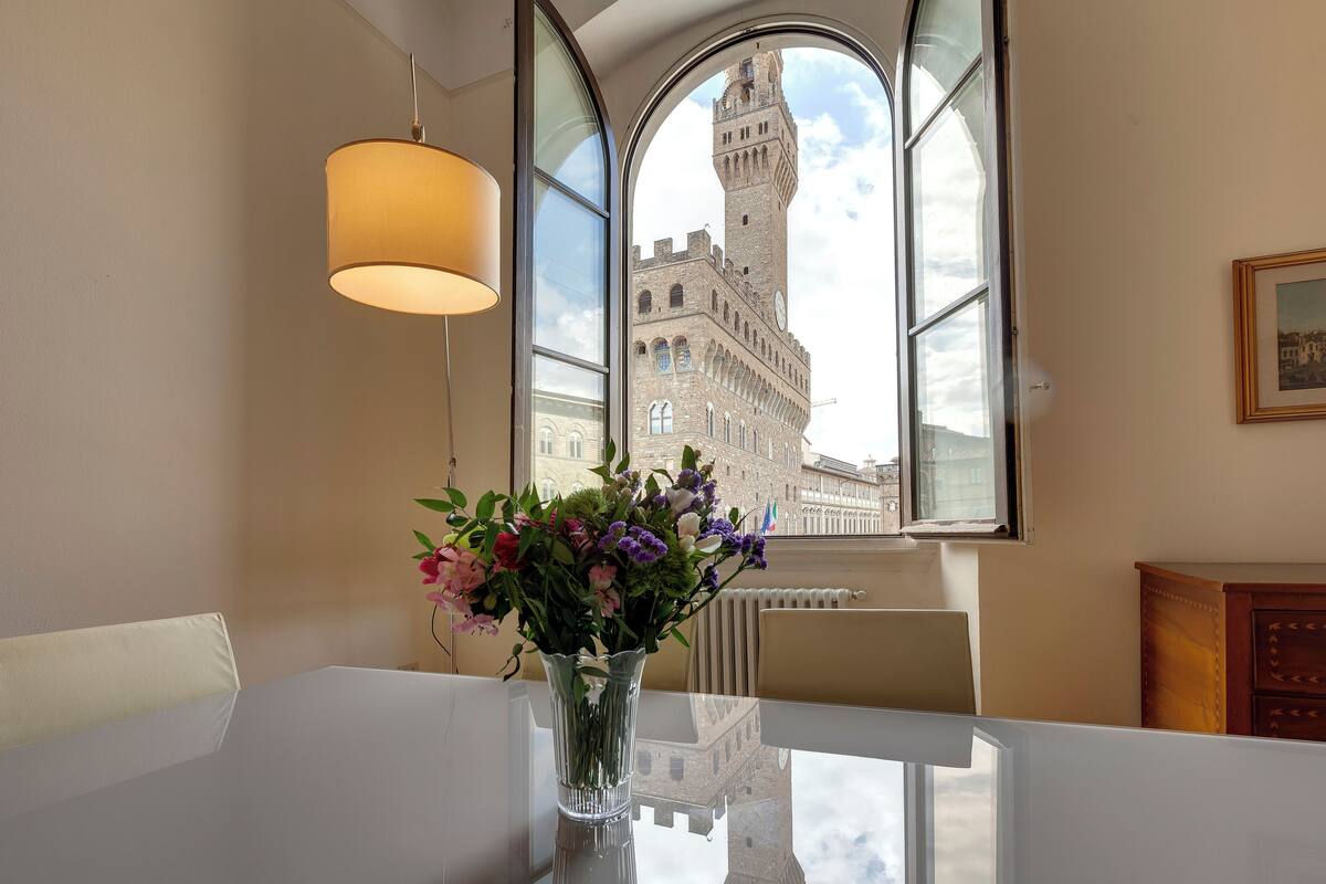 A room with a dining table, a vase of flowers, a lamp, and a view of a historic building through a window.