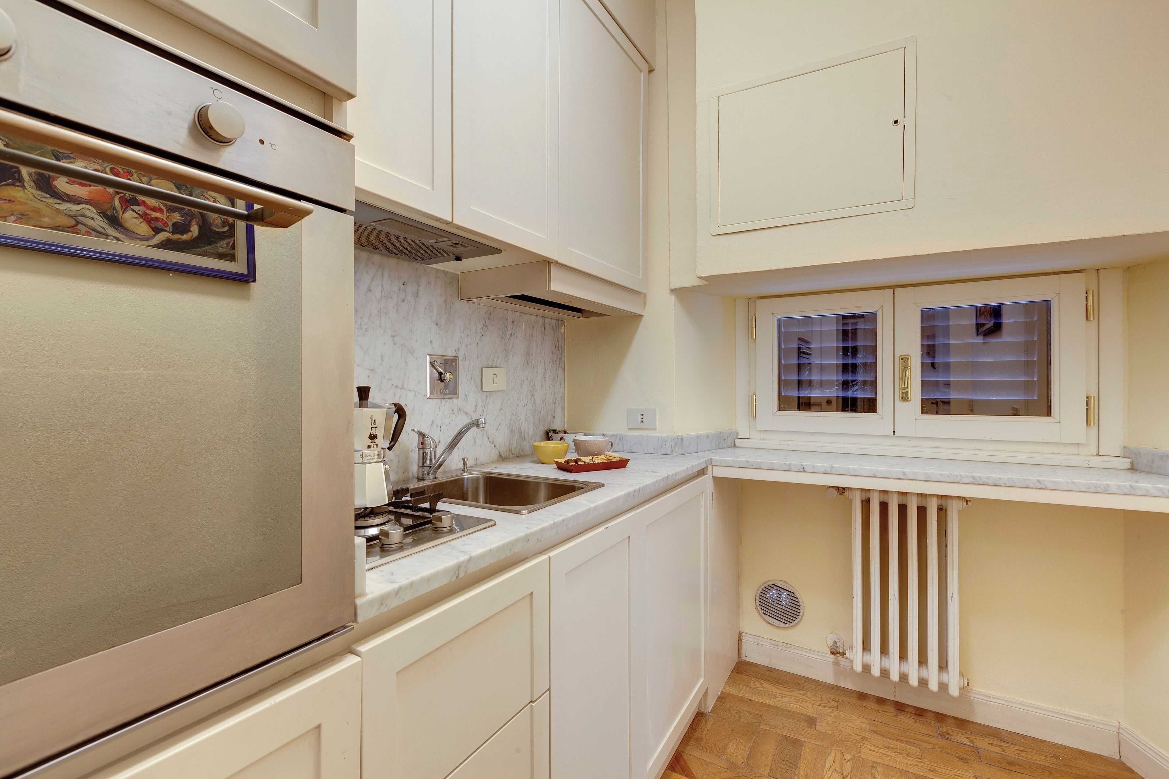 Una cocina con armarios blancos, un refrigerador de acero inoxidable, un salpicadero de mármol, una ventana con persianas y un radiador.