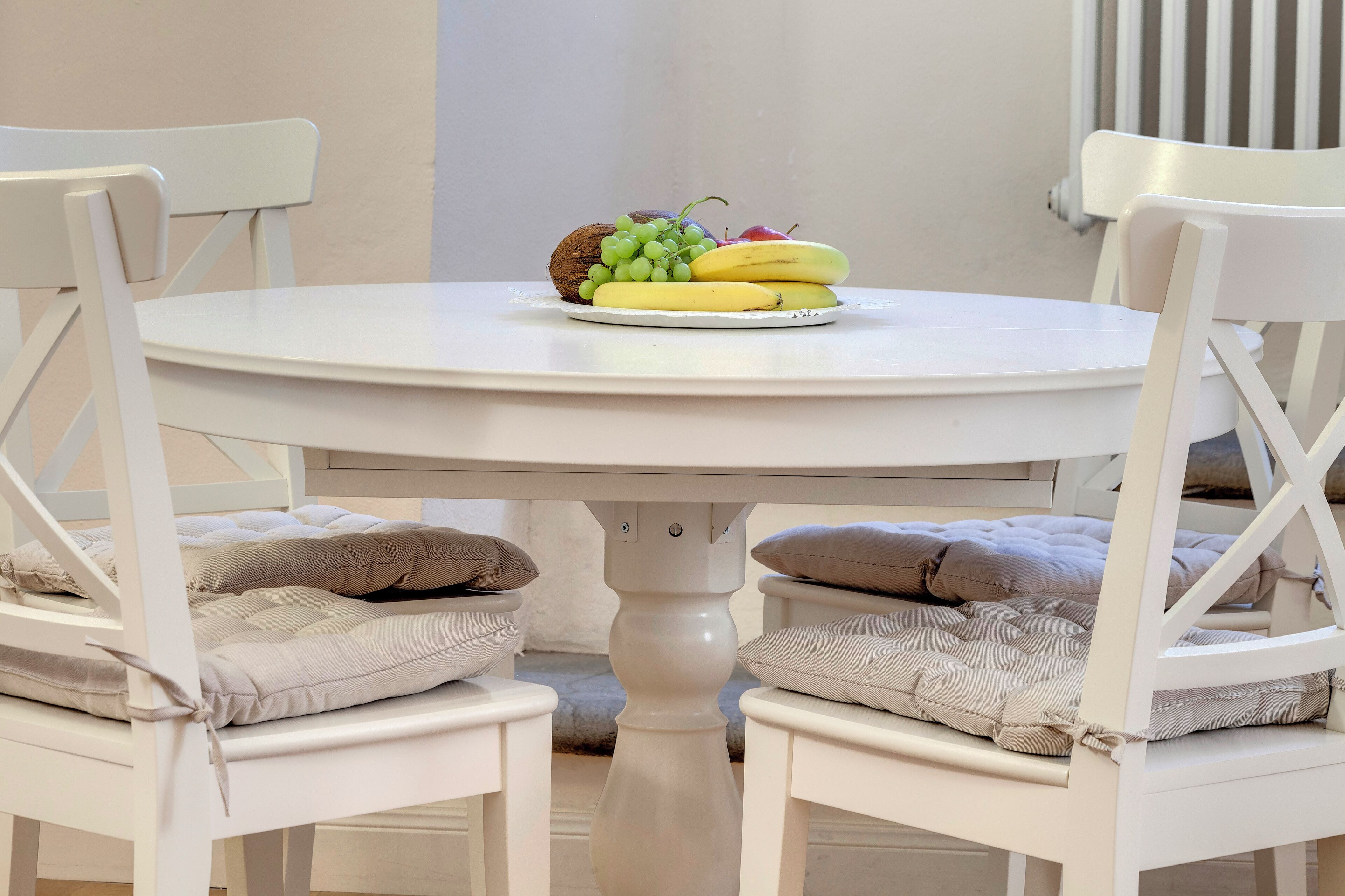 A round white table with a fruit plate, two white chairs with cushions, and a white wall with a radiator.