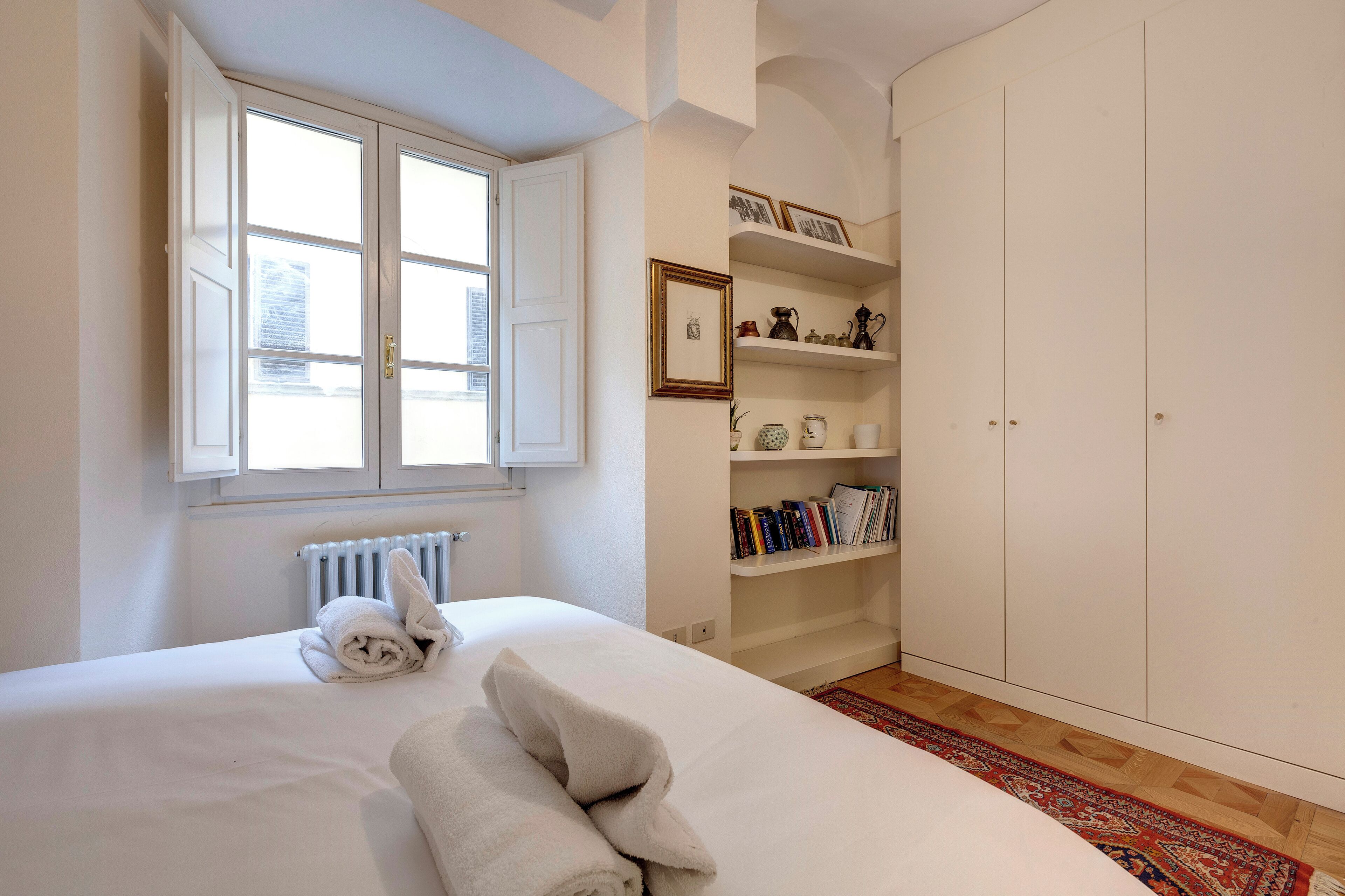 Une chambre à coucher comprenant un lit, une fenêtre, un radiateur, une armoire et des étagères avec des livres et des objets décoratifs.
