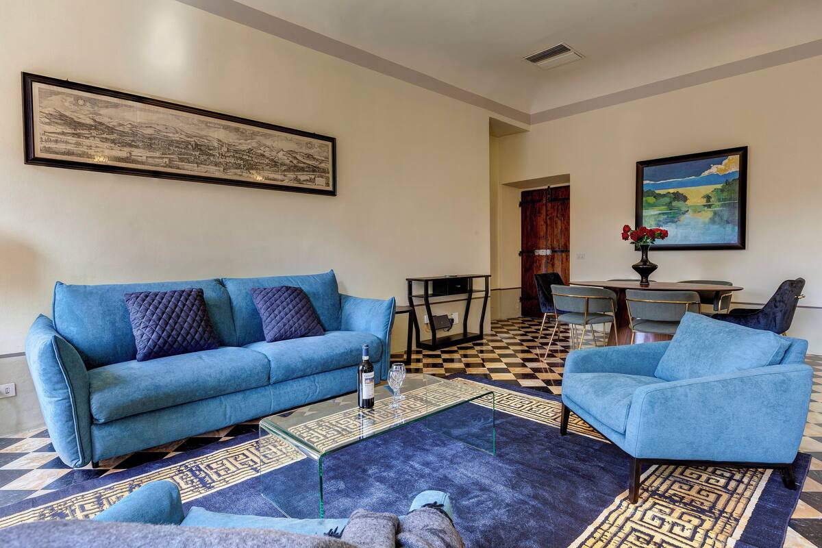 A living room with a blue sofa, a coffee table, and a patterned rug. There is a dining table with chairs in the background.