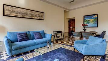 Ein Wohnzimmer mit einem blauen Sofa, einem Couchtisch und einem Teppich mit Muster. Im Hintergrund steht ein Esstisch mit Stühlen.