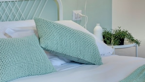 A neatly made bed with green knitted pillows, a white headboard, and a bedside table with a plant and a bottle. - Mamo Florence - Maddalena Apartment (Florence)
