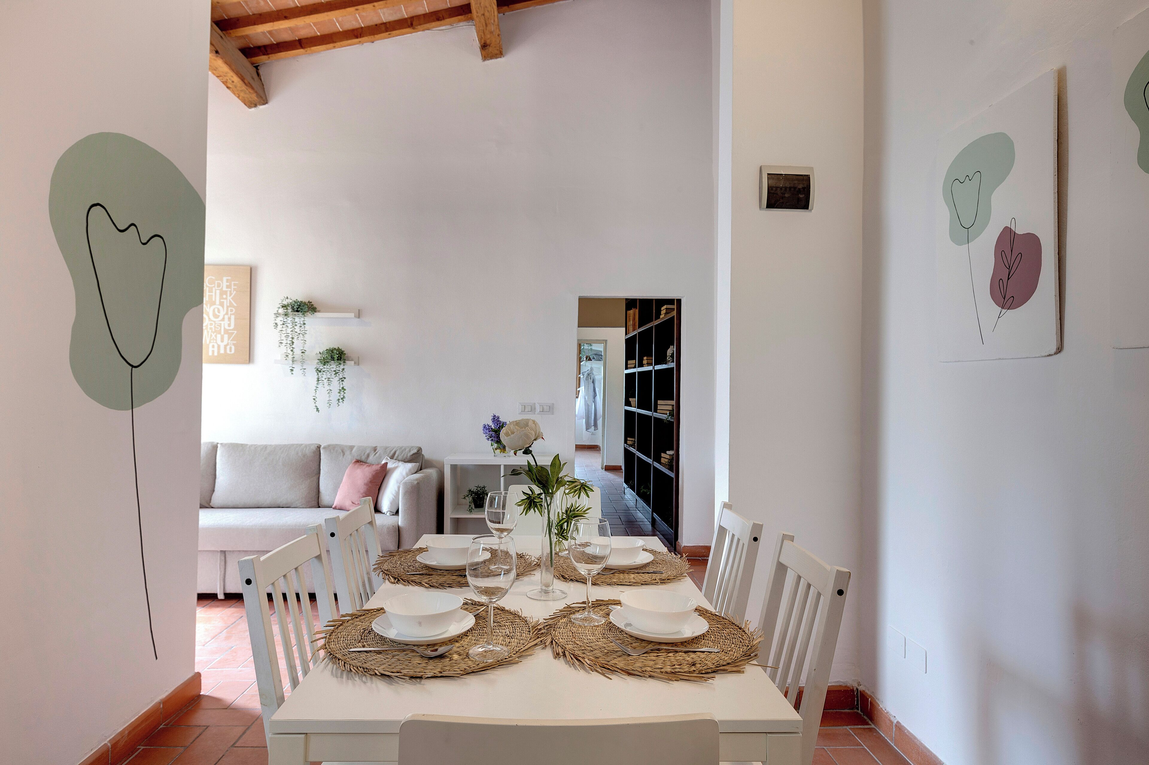 A dining area with a white table set for four, surrounded by white chairs, with a wooden ceiling and a bookshelf in the background.