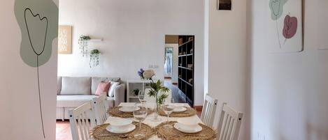 A dining area with a white table set for four, surrounded by white chairs, with a wooden ceiling and a bookshelf in the background.