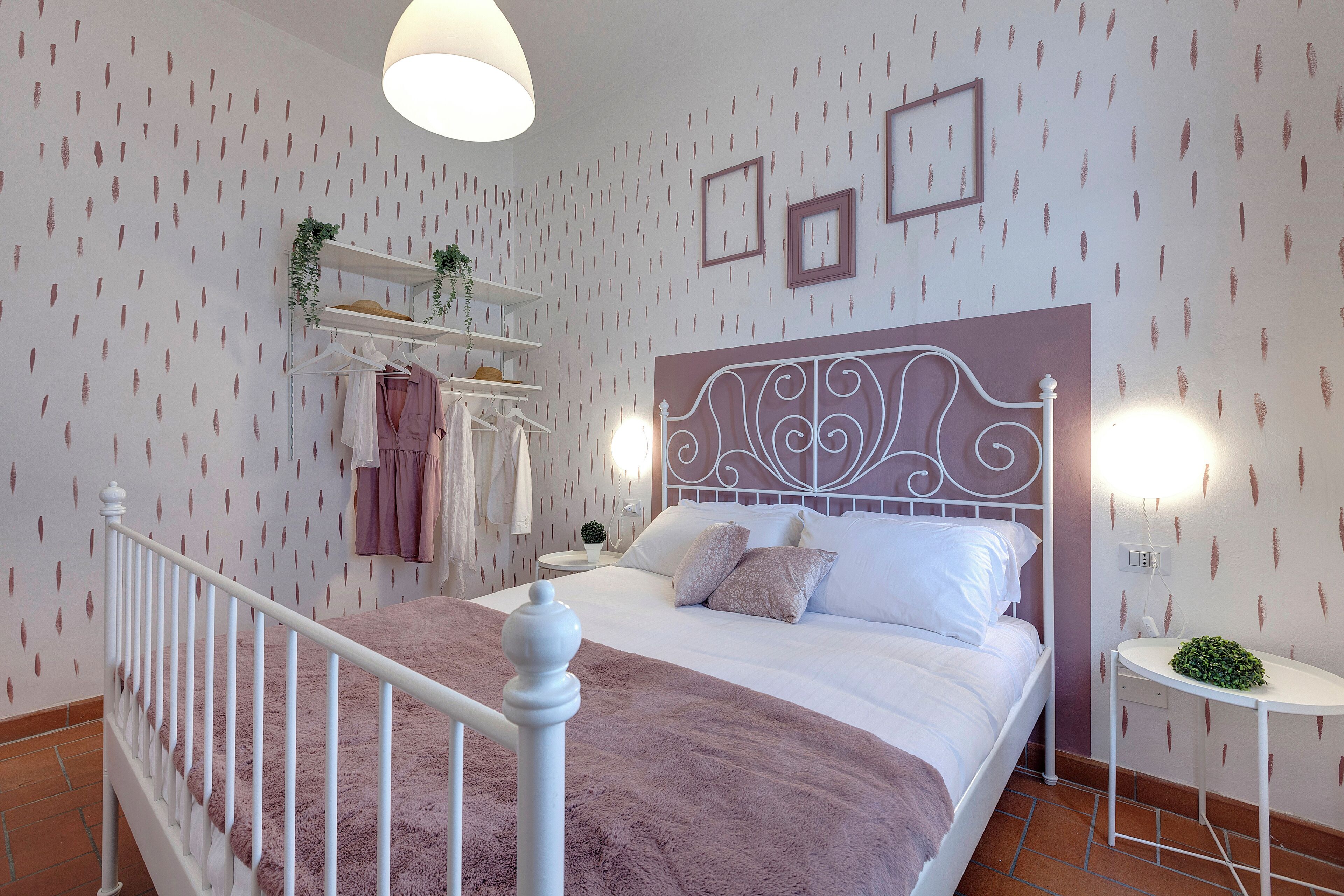 A bedroom with a bed, white metal headboard, bedside table, and wall-mounted shelves with clothes and plants.