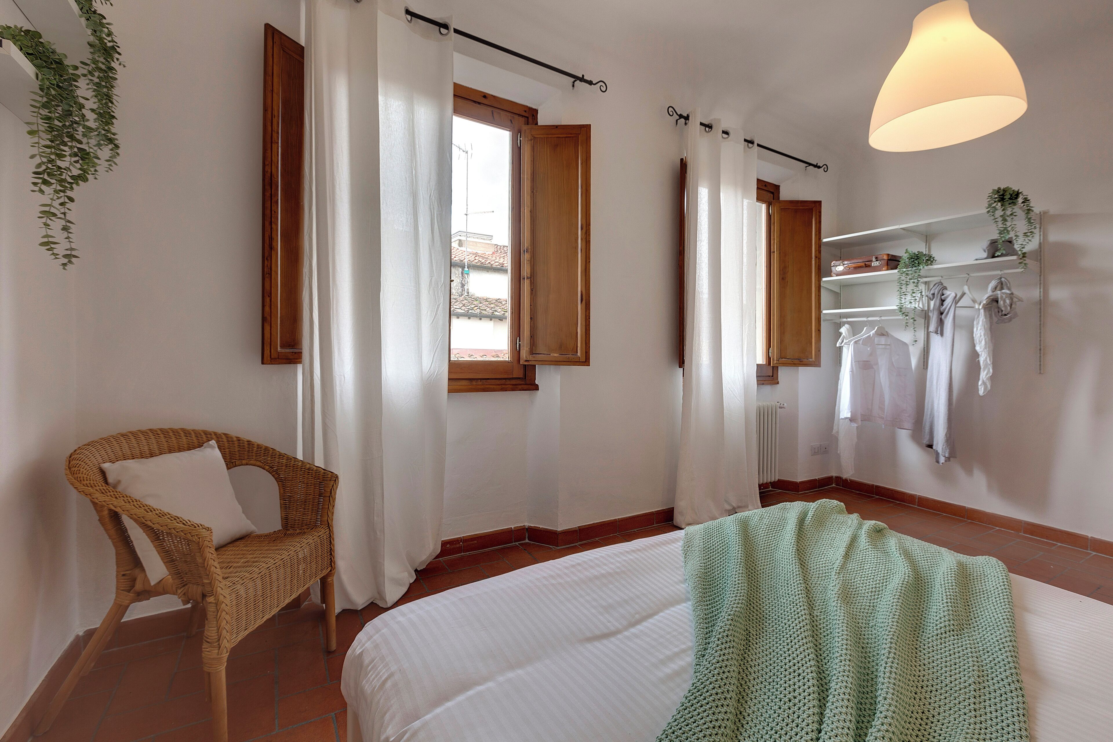 A bedroom with a bed, a wicker chair, a window with curtains, a hanging plant, and a shelf with clothes.