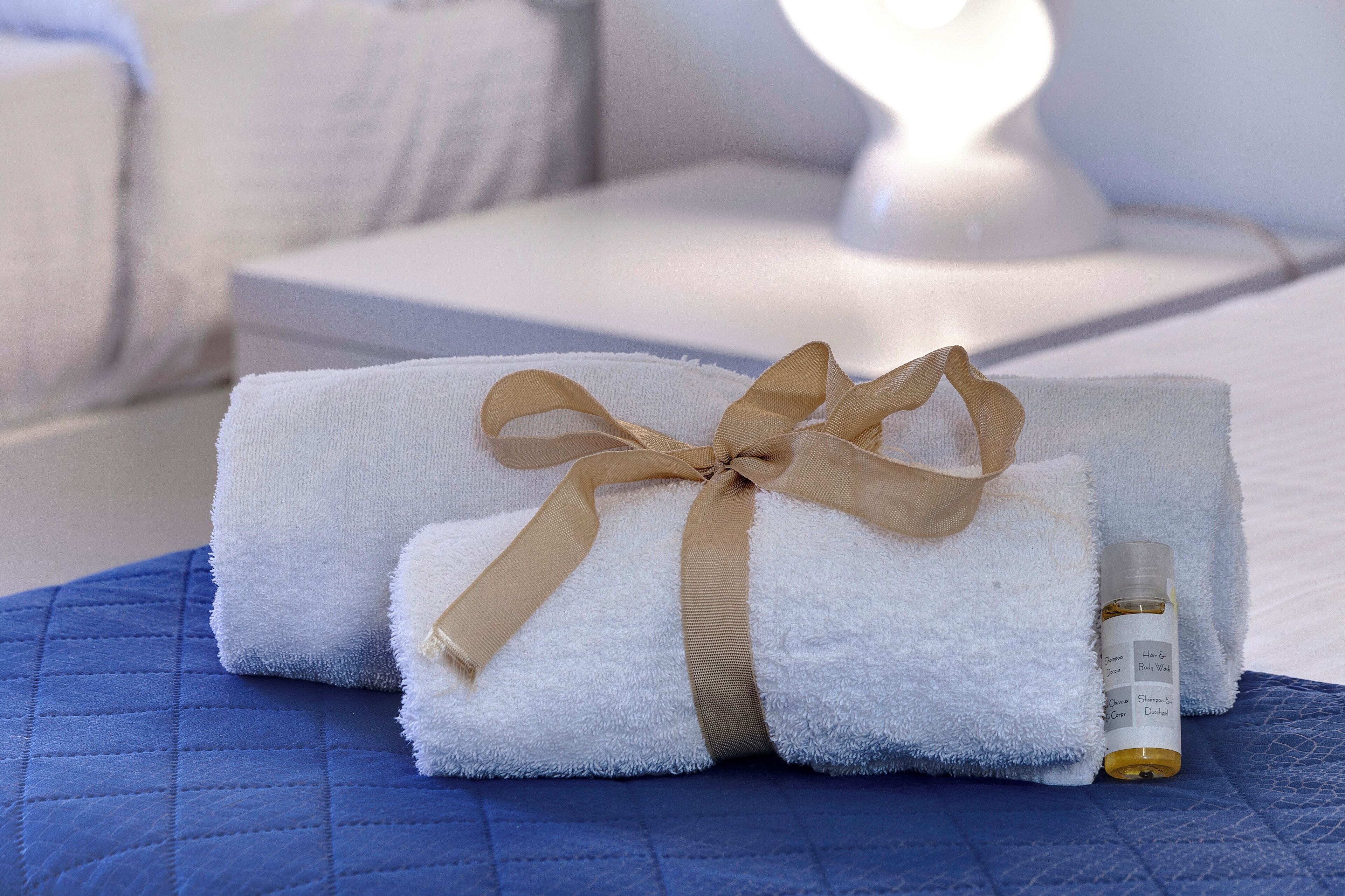 A neatly folded white towel with a beige ribbon tied around it, placed on a blue quilted bedspread.