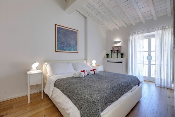 A neatly made bed with a gray blanket, white pillows, and two rolled towels with red ribbons. There is a bedside table with a lamp, a wall-mounted painting, and a window with sheer curtains.