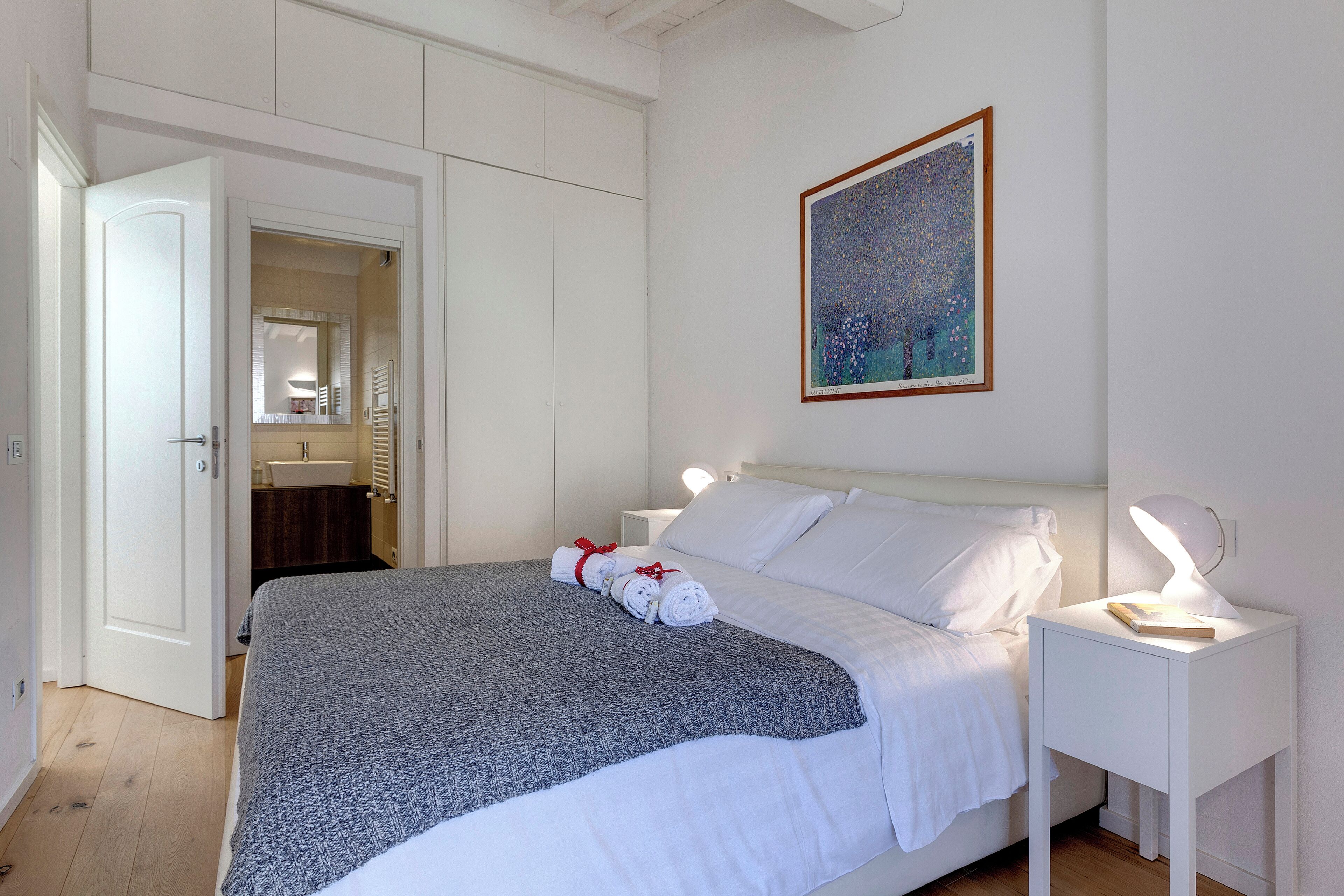 Ein ordentlich gemachtes Bett mit weißen Leinen und einer grauen Decke, ein Nachttisch mit einer Lampe und ein Gemälde an der Wand.