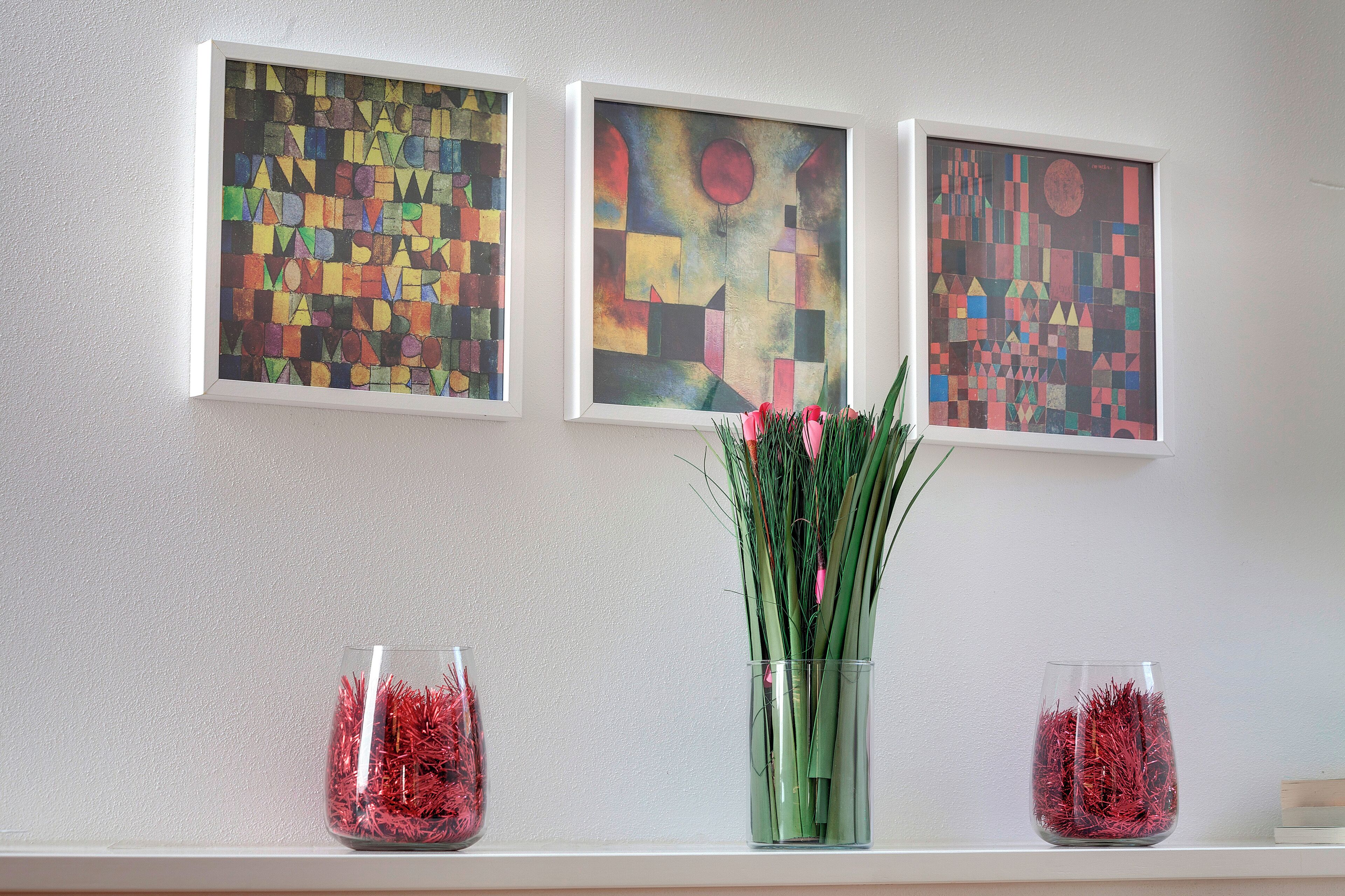 Three framed abstract paintings on a wall, a vase with green plants in the center, and two glass vases with red decorative items on a shelf.