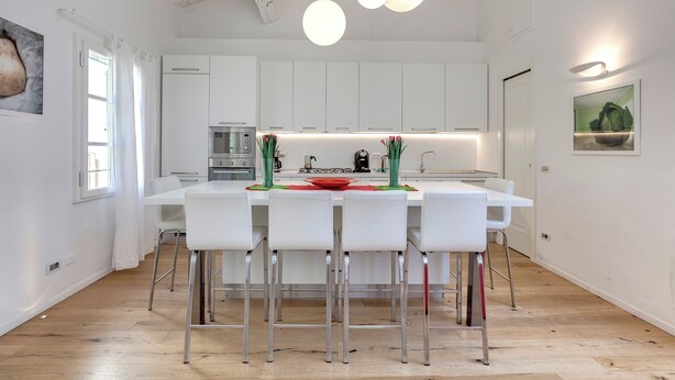 A modern kitchen with white cabinets, a central island with bar stools, pendant lighting, and a view of the dining area. - Mamo Florence - Margot Apartment (Florence)