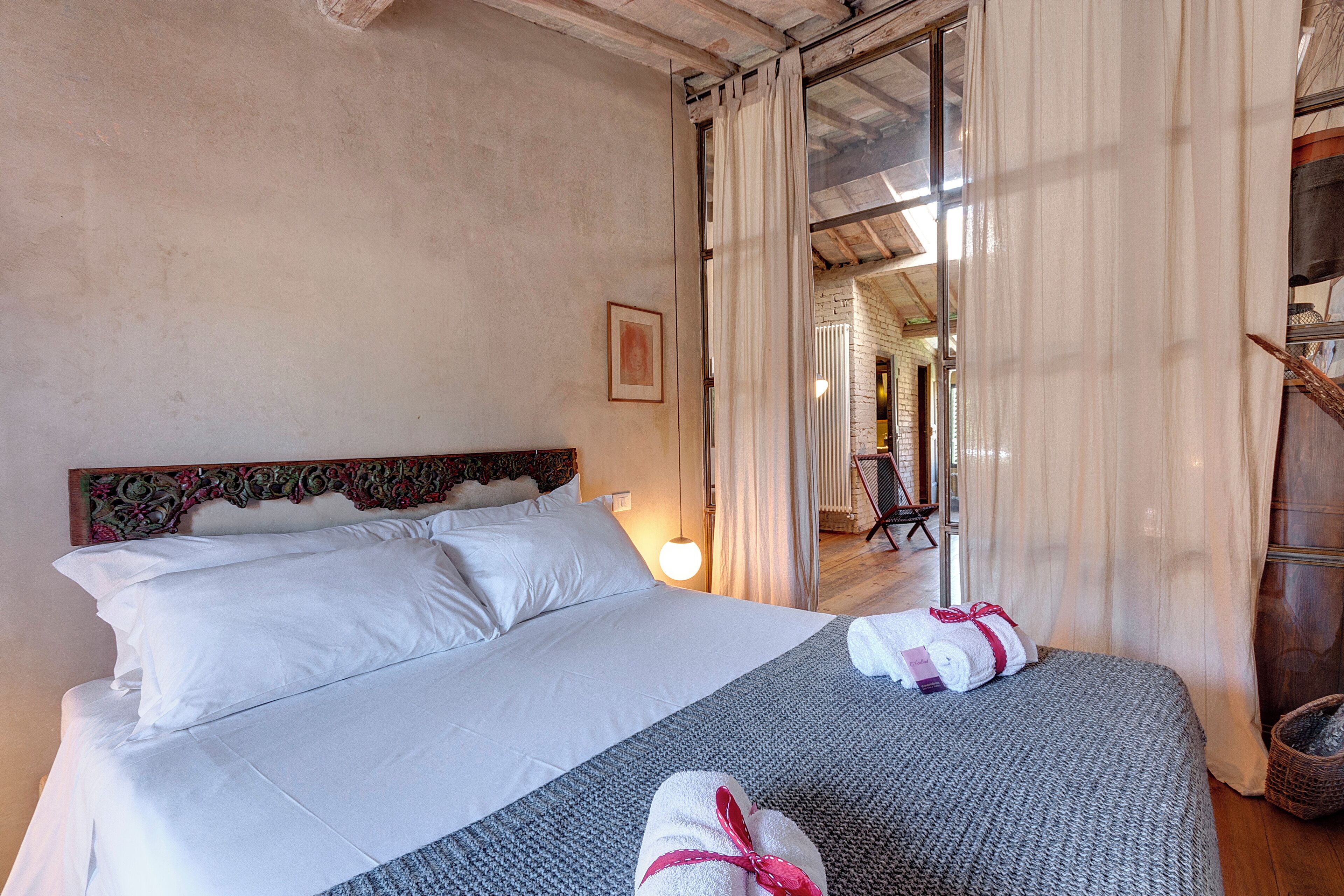 A hotel room with a large bed, white linens, a headboard with a floral pattern, a framed picture on the wall, and a window with sheer curtains.