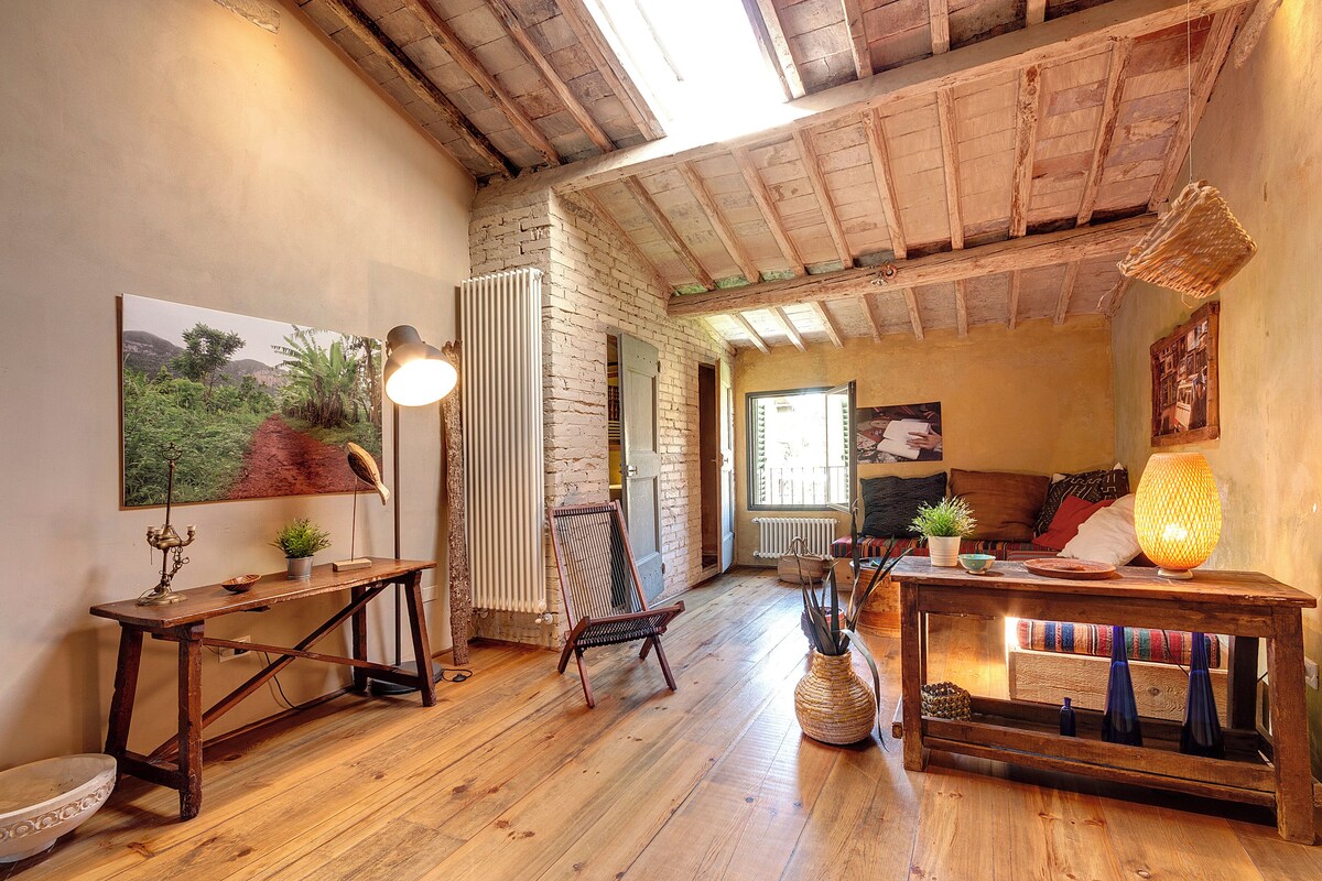A rustic room with exposed wooden beams, a wooden floor, a table with a lamp, a chair, a radiator, and a painting on the wall.
