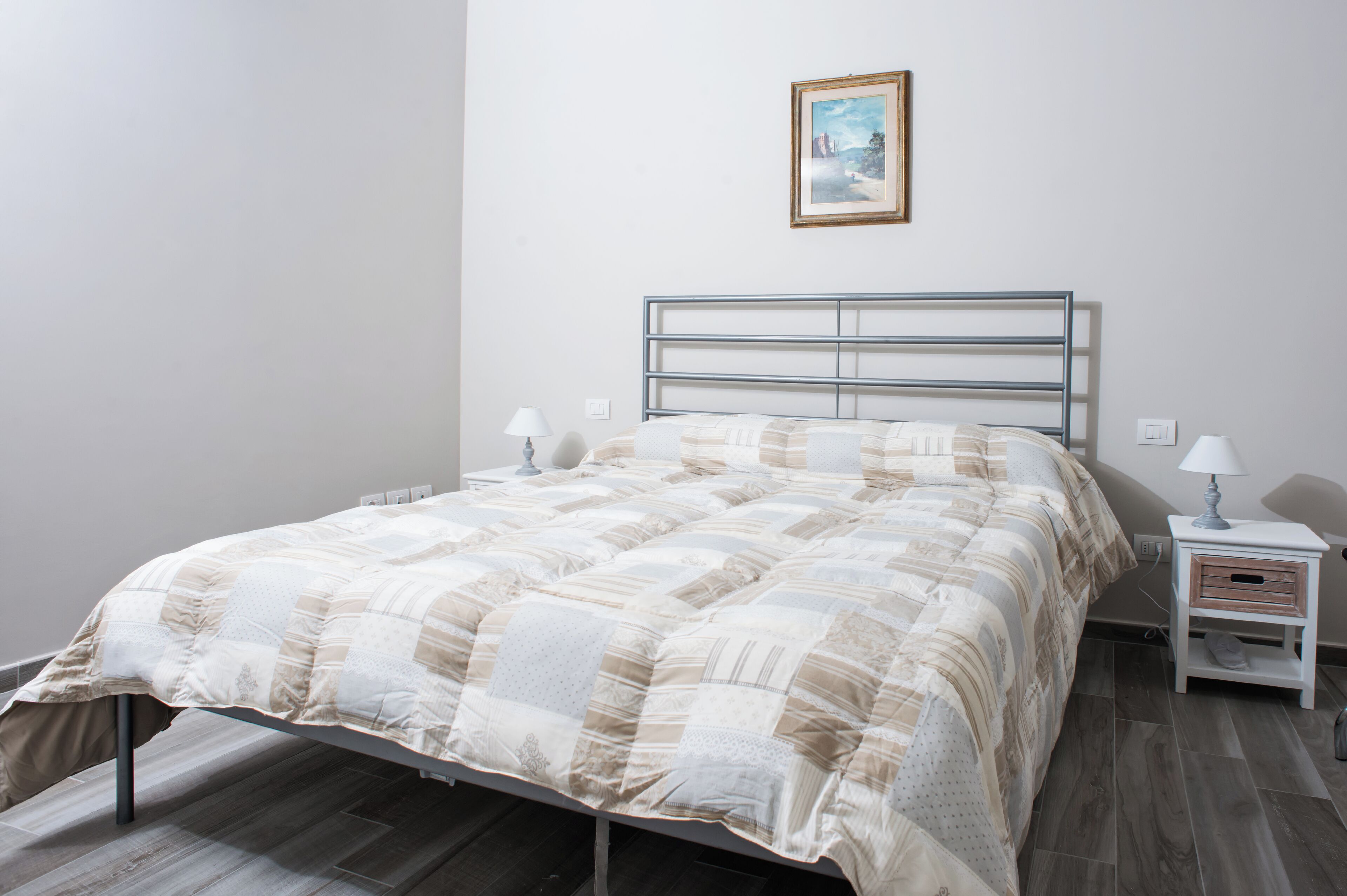 A neatly made bed with a metal frame, a bedside table with a lamp, and a small drawer unit.