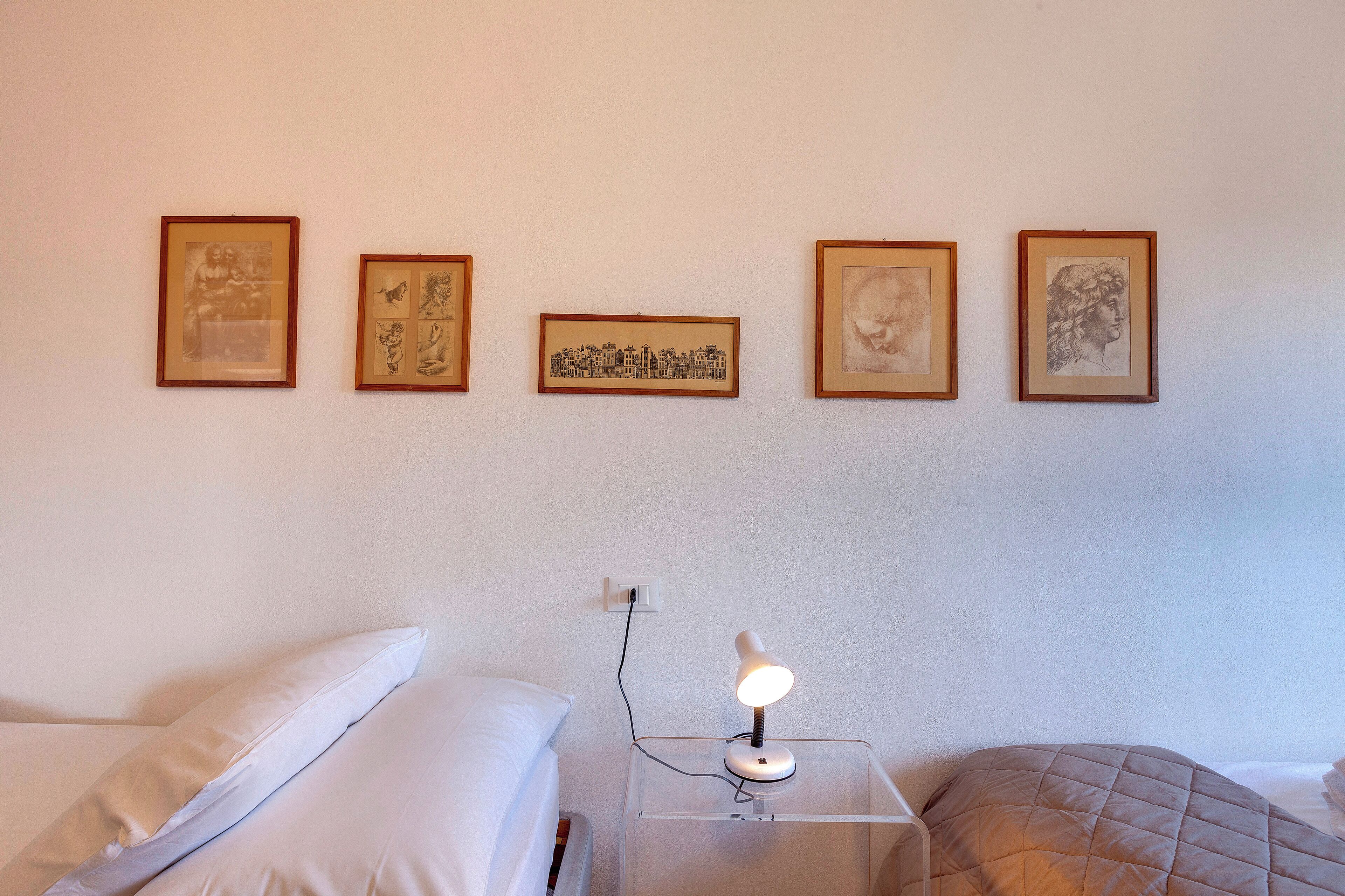 Un dormitorio con cama, una mesita de noche con lámpara y cuatro obras de arte enmarcadas en la pared.