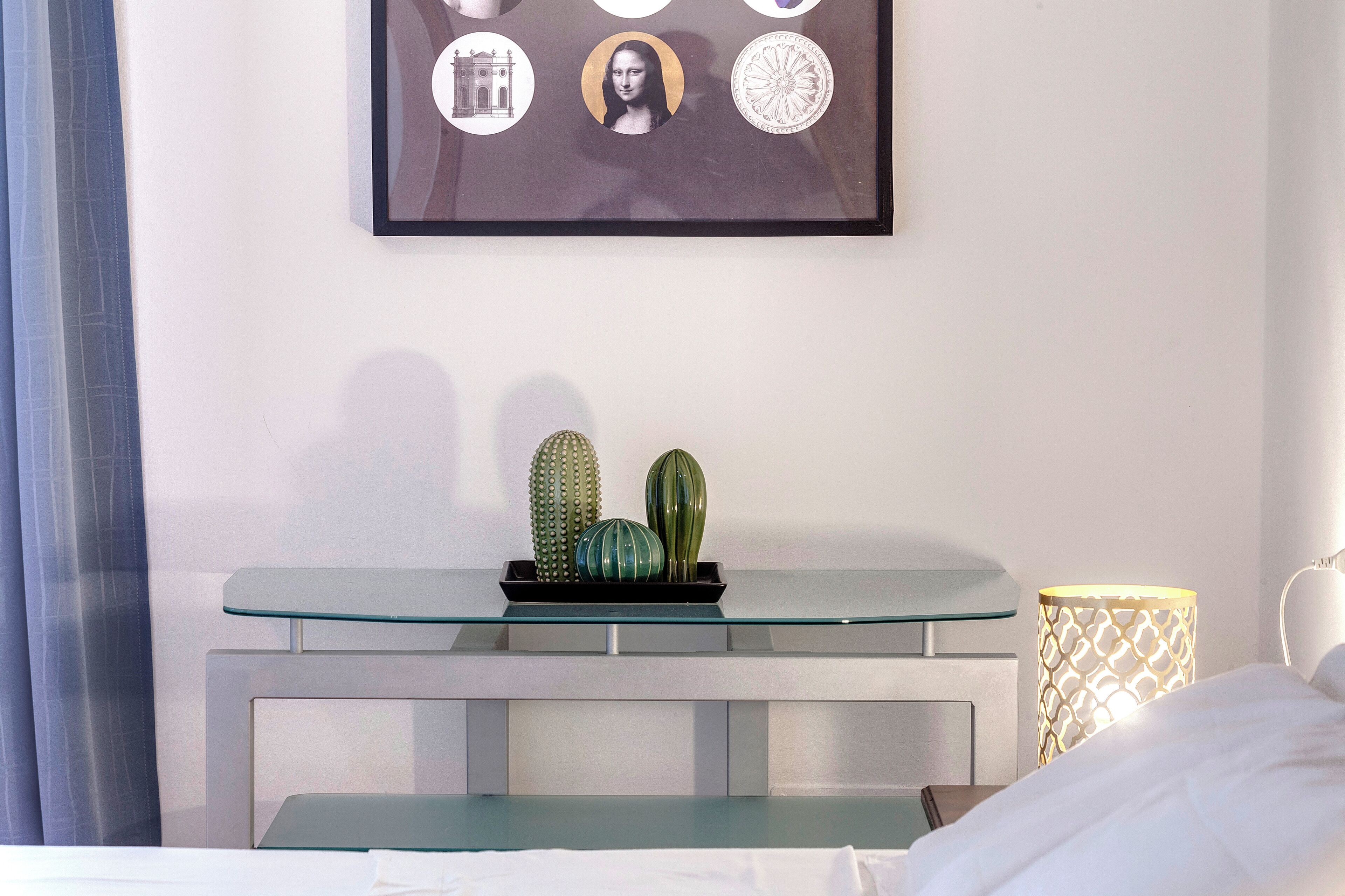 A hotel room with a glass-top desk, a bed with white linens, a framed artwork above the desk, and a decorative lamp on the side.