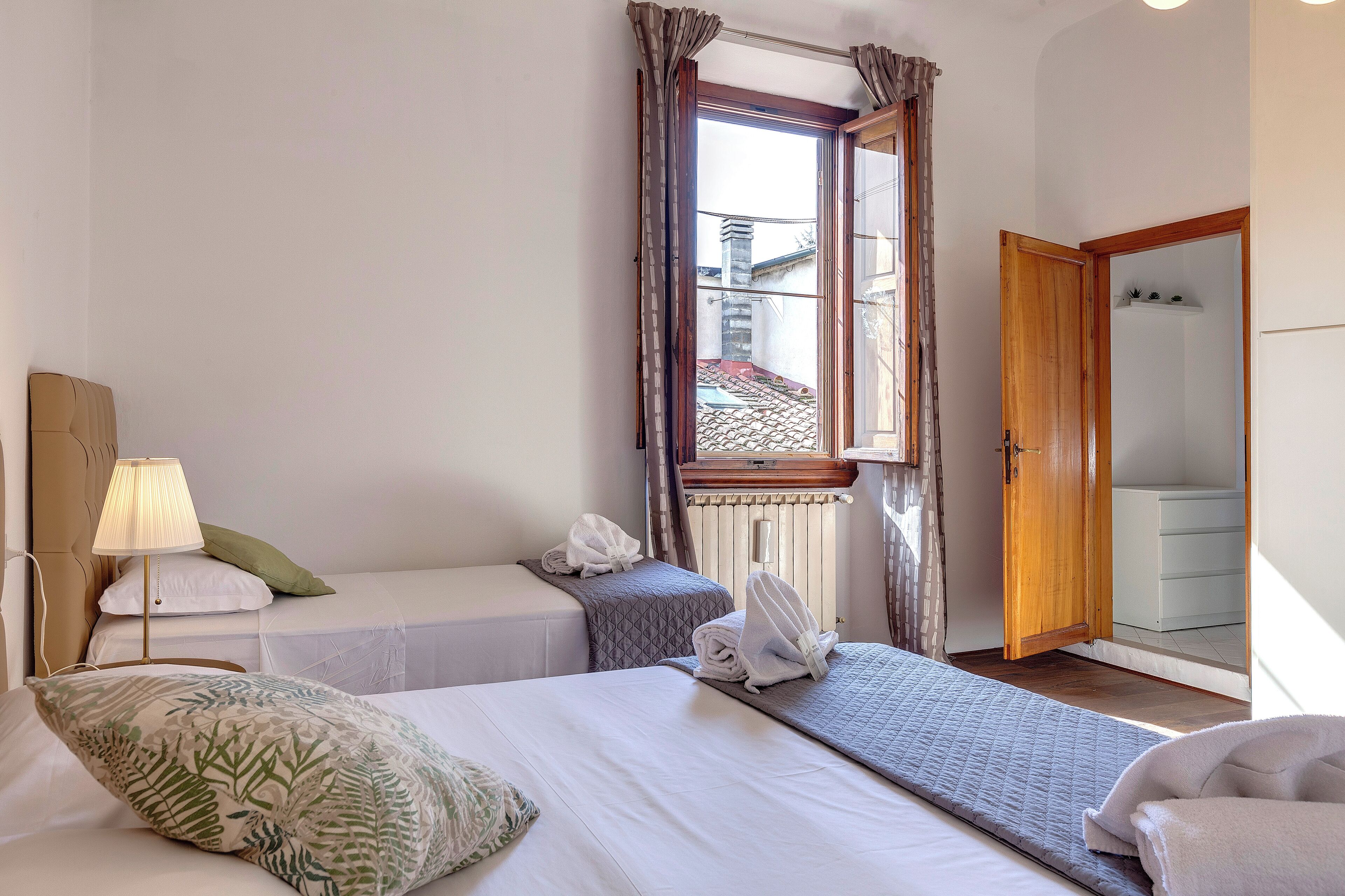 Une chambre d’hôtel avec deux lits, une fenêtre avec des rideaux, une lampe et une porte en bois donnant sur une salle de bain.