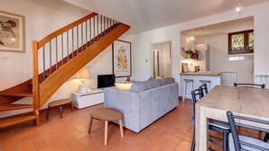 A living room with a wooden staircase, a grey sofa, a wooden dining table, and a kitchen area with a window.
