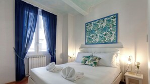 A neatly made bed with white linens and towels, a blue curtain, a wall-mounted artwork, and a bedside table with a lamp and a vase. - Mamo Florence - San Lorenzo Apartment (Florence)