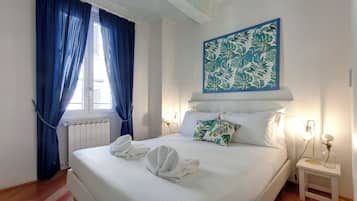 Cama bem arrumada com lençóis e toalhas brancos, cortina azul, obra de arte na parede e mesa de cabeceira com abajur e um vaso.