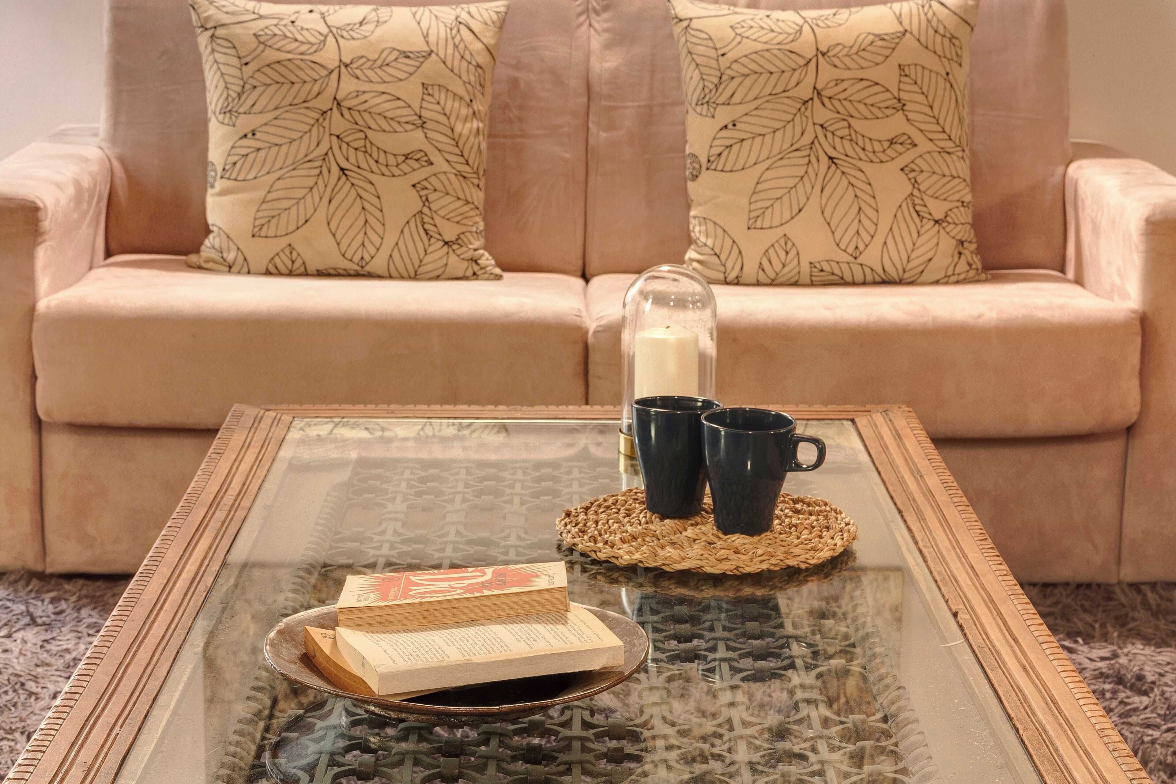 Une table basse avec un plateau en verre, sur laquelle reposent un set de table en fibres tressées, deux tasses noires et une pile de livres.