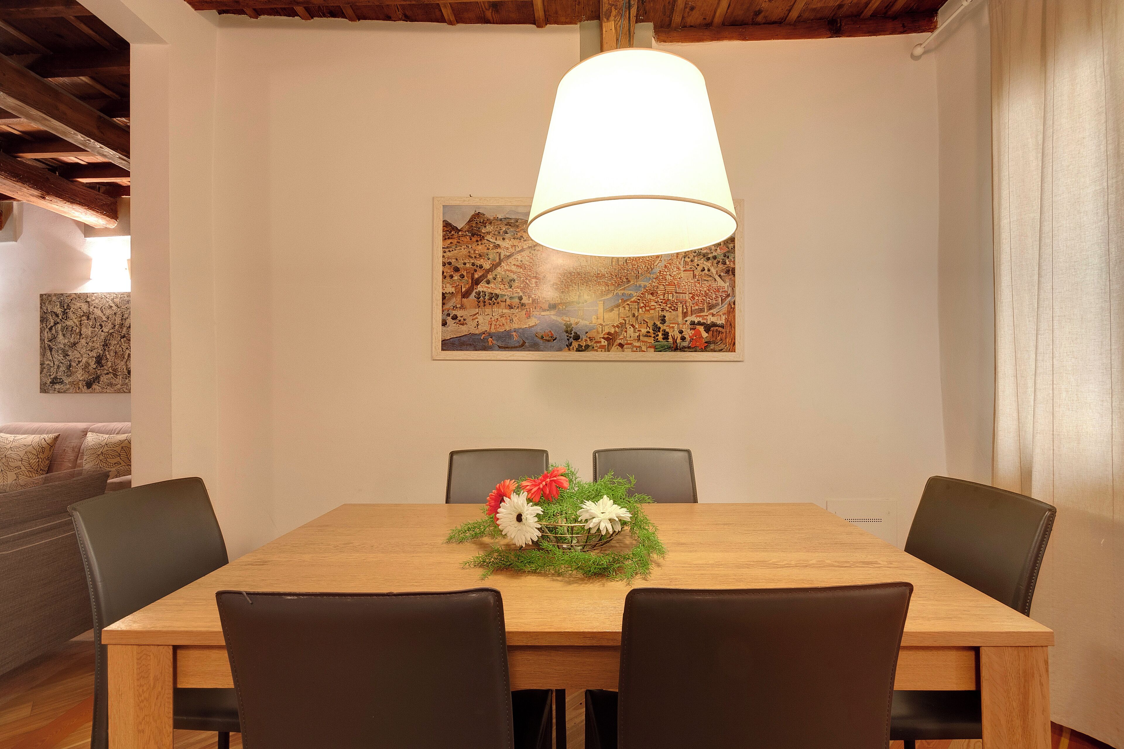 Zona pranzo con tavolo in legno, sedie nere, un centrotavola floreale e un quadro appeso al muro.