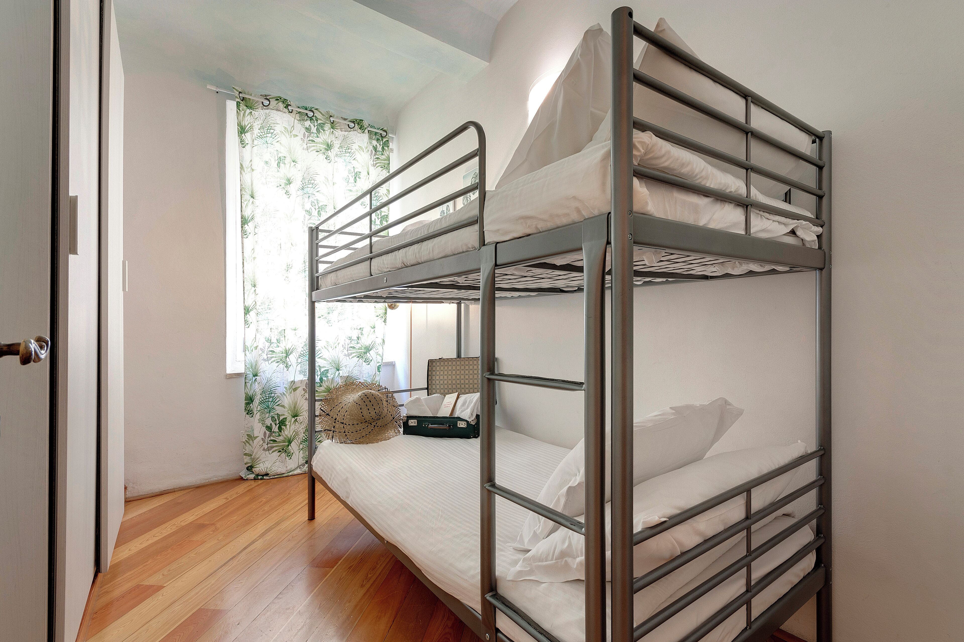Quarto com beliche, chão de madeira, janela com cortinas e uma mala no beliche inferior.