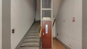 A narrow stairway with a wooden handrail, a red door at the bottom, and a window with a mesh screen above.