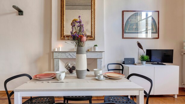 A dining area with a table set for two, a vase with flowers, a fireplace, a television, and a cabinet.