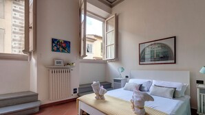 A bedroom with a bed, a radiator, a window with curtains, a framed picture, and a small table with a lamp.