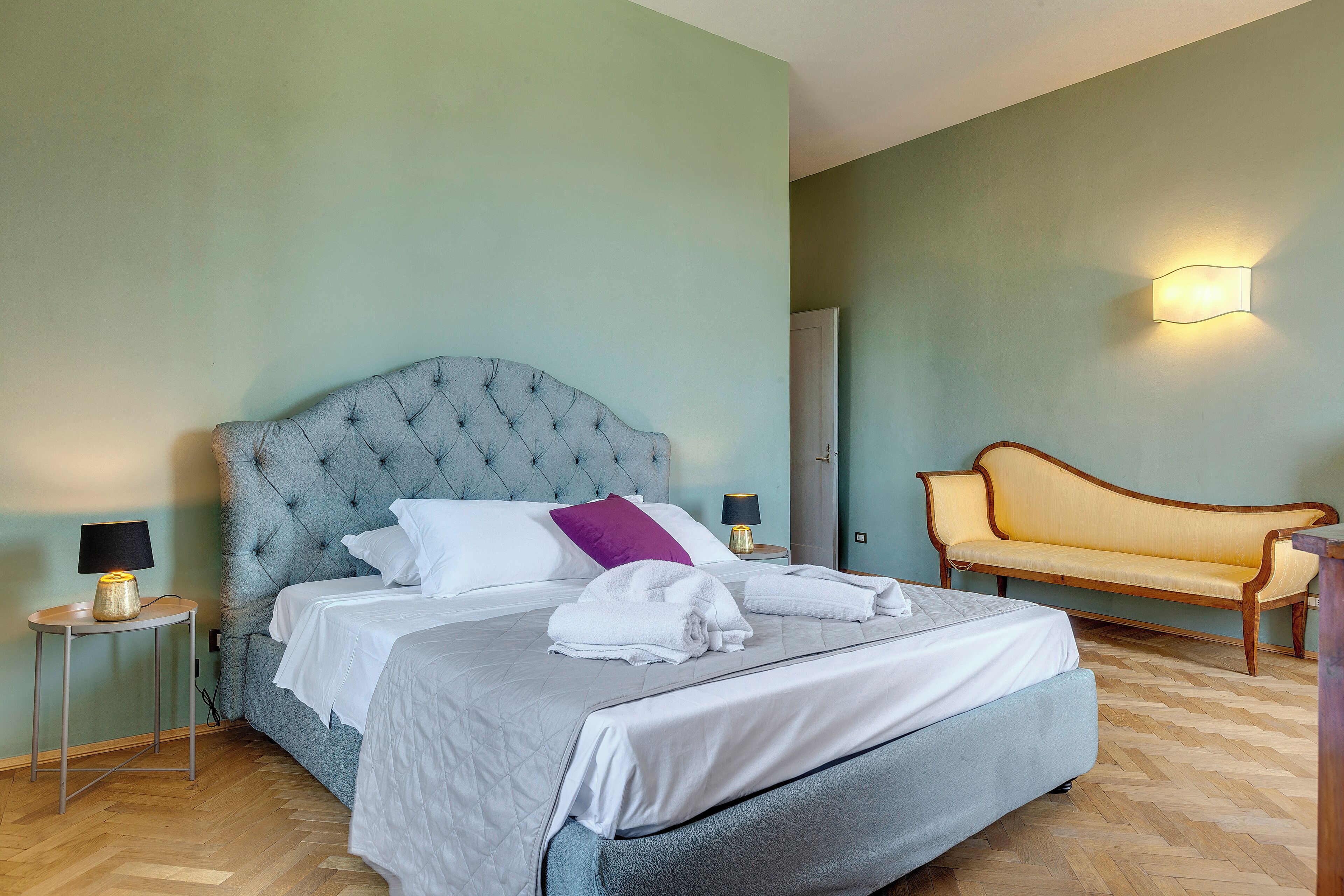 Ein ordentlich bezogenes Bett mit einem gebogenen Kopfteil, ein Nachttisch mit Lampe sowie ein Sofa in einem Zimmer mit hellgrünen Wänden und Holzdielen.