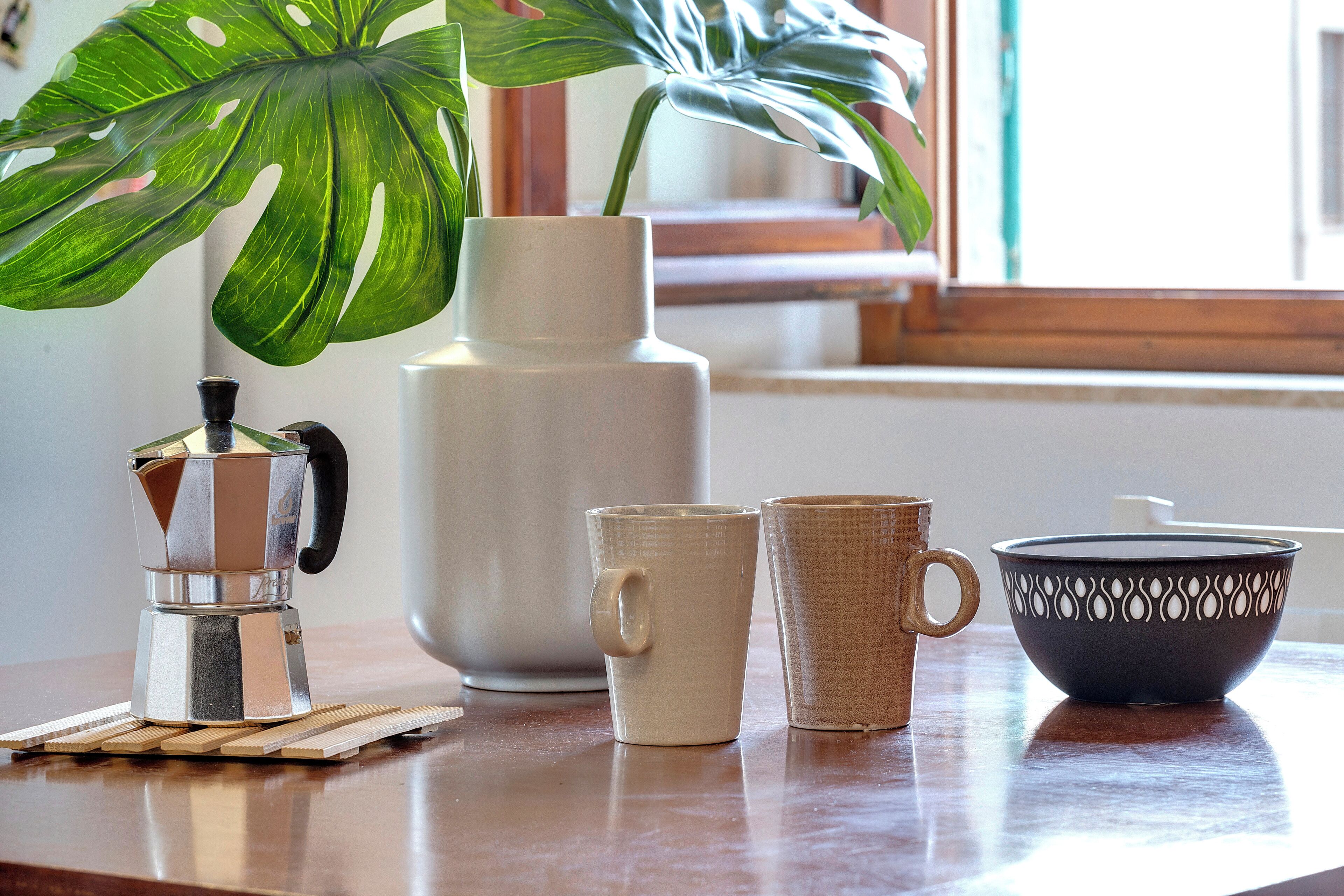 Ein Holztisch mit einer Kaffeemaschine, zwei Tassen, einer Schüssel und einer Blumentopf-Pflanze.