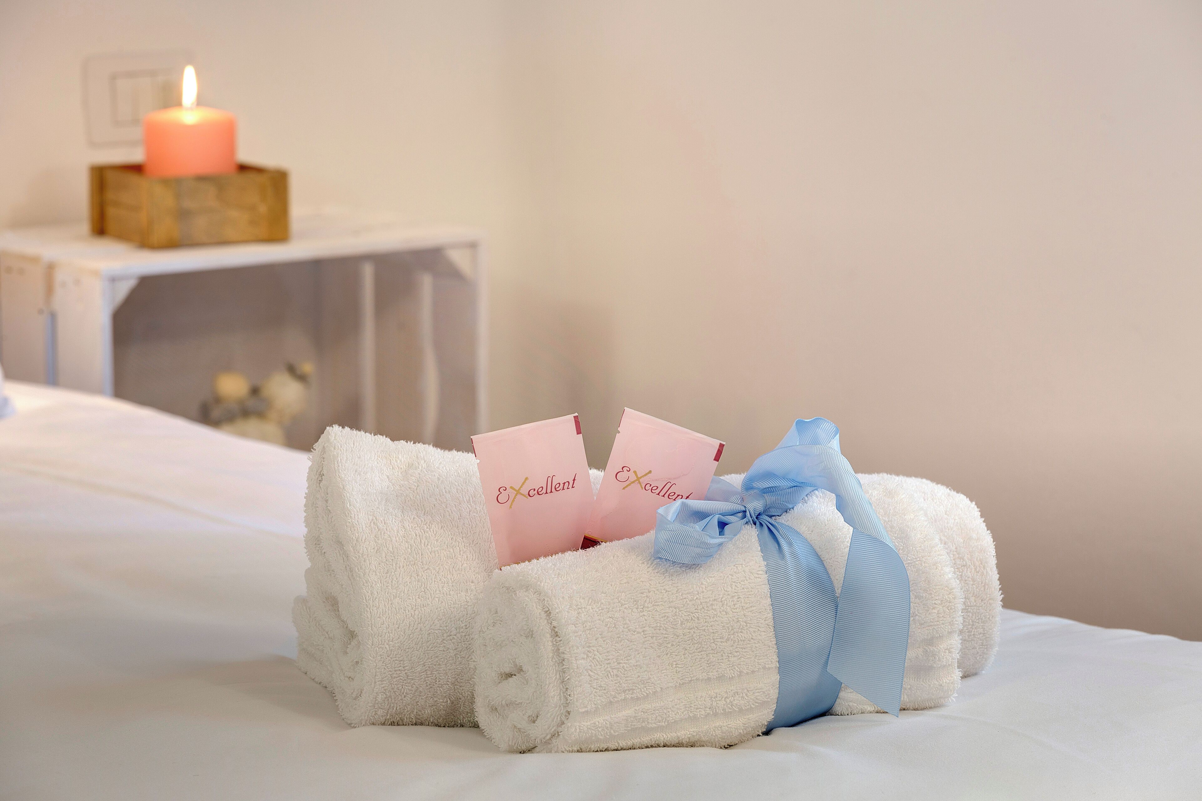 A neatly made bed with a rolled white towel, a lit candle, and two pink cards with the word "Excellent" on them.