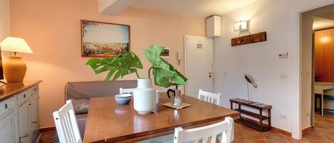 Een eethoek met een houten tafel, witte stoelen, een vaas met een plant, een lamp en een schilderij aan de muur.