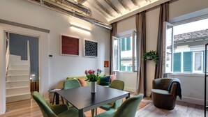 Una sala moderna con una mesa de comedor, sillas verdes, un sofá con cojines coloridos, una escalera y un ventanal con vistas a edificios.