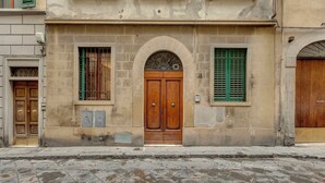 Un edificio con un arco central, flanqueado por dos ventanas con contraventanas verdes, y una calle empedrada en el frente.
