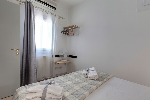 Un dormitorio con cama, ventilador de cabecera, toallero y ventana con cortinas.