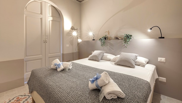 A neatly made bed with a grey blanket and towels, a bedside lamp, and a wall-mounted shelf with decorative items. - Mamo Florence - Zante Aparment (Florence)
